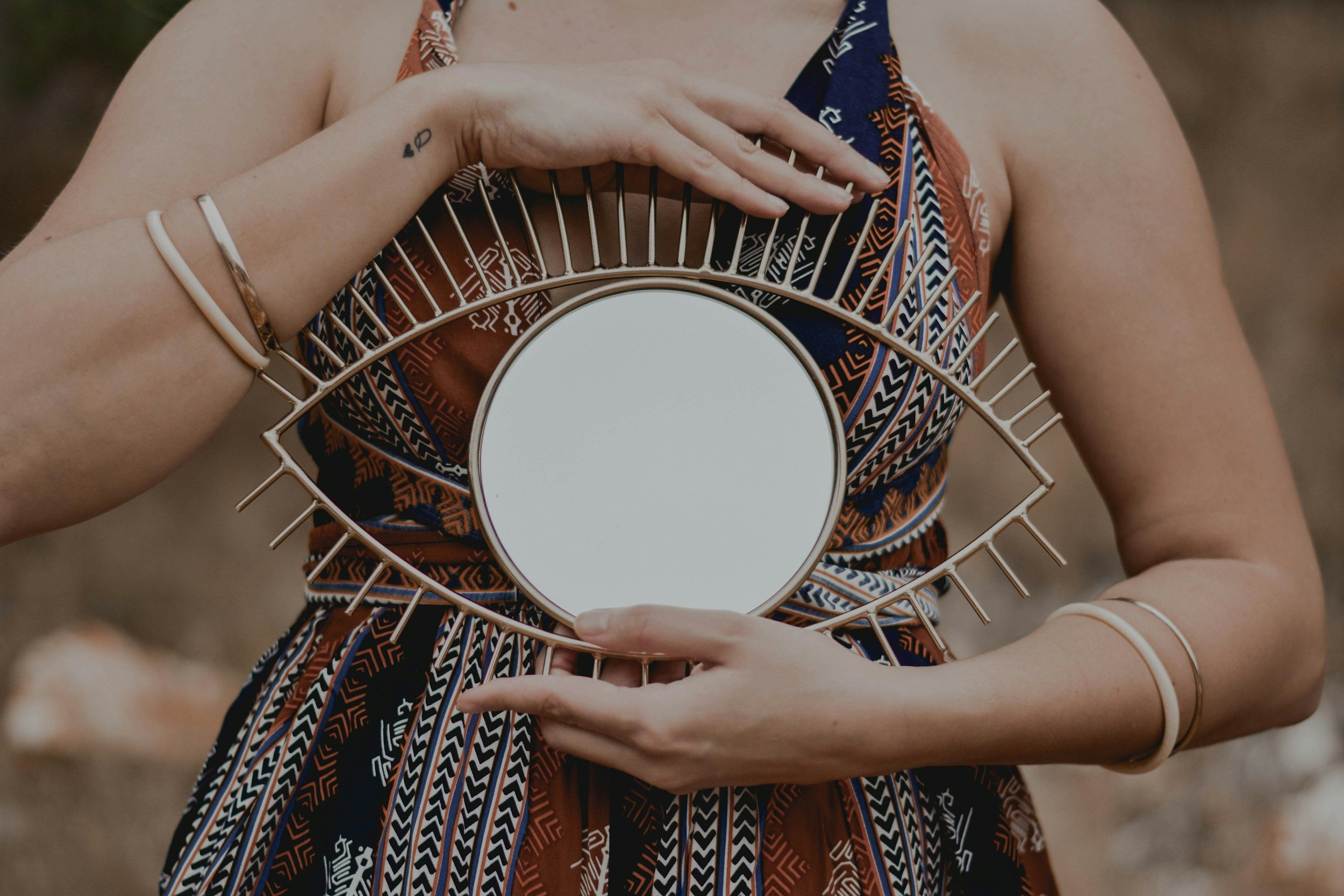 A woman holding a decorative mirror with an eye-shaped frame featuring gold spokes and a circular reflective surface, wearing a colorful patterned dress and silver jewelry.