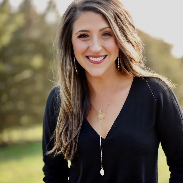 A smiling woman with long, wavy brown hair wearing a black top and jewelry outdoors.