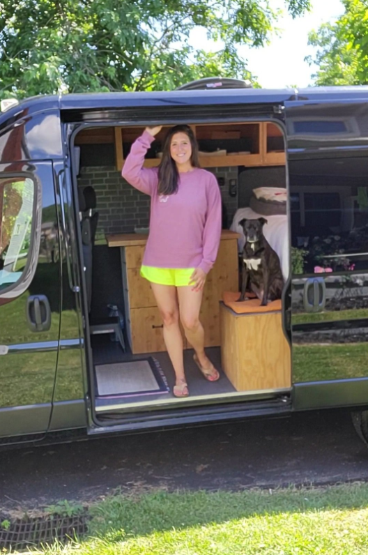 A smiling young woman in a pink sweater and yellow shorts stands inside a converted van, waving. A dog is sitting on a wooden platform next to her, with the van's interior visible, including bed and storage areas.