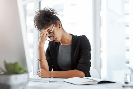 A woman sitting at a desk with her hand on her forehead, looking down at an open notebook, in a bright office setting.