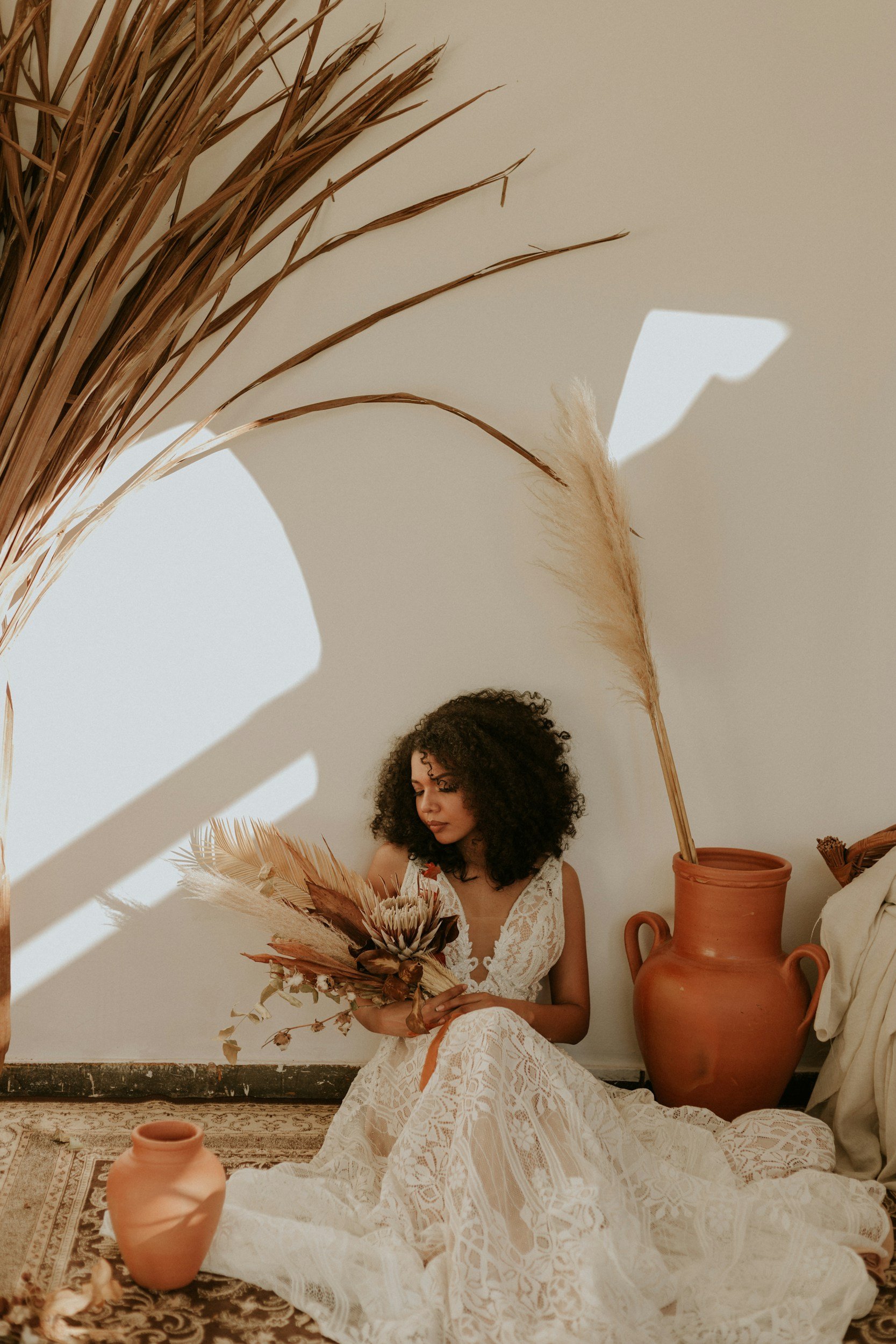 A woman with curly hair sits on a patterned rug, holding a bouquet of dried flowers, surrounded by large terracotta vases and dried plants in a minimalistic setting.