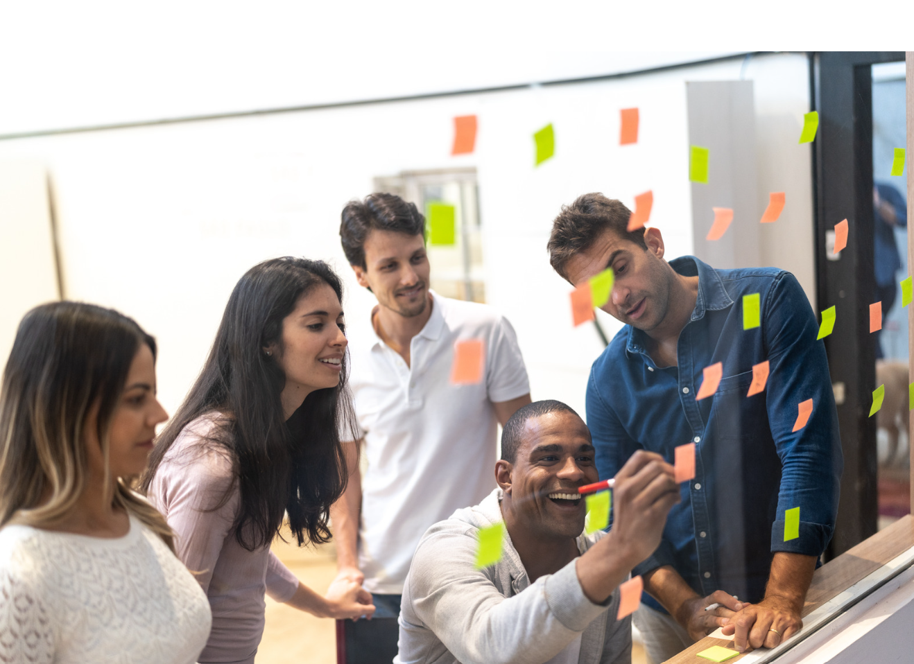 Group of five diverse young adults collaborating and brainstorming, placing colorful sticky notes on a glass wall in an office.