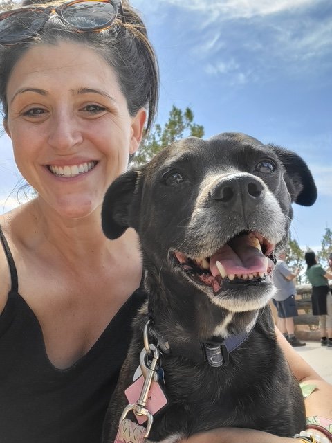 A woman smiling with sunglasses on her head poses with a happy black dog outside on a sunny day.