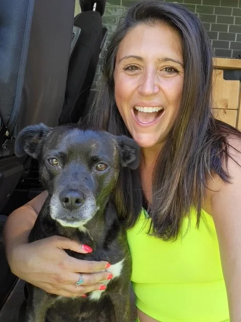 A woman with long dark hair taking a selfie with a black dog with white markings on its face, both smiling. The woman is wearing a bright yellow sleeveless top and is outdoors.
