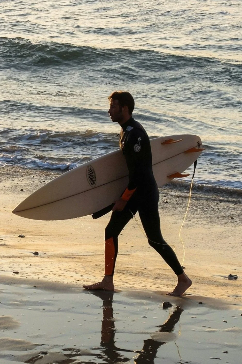 A man in a wetsuit walking along the beach carrying a surfboard at sunset.