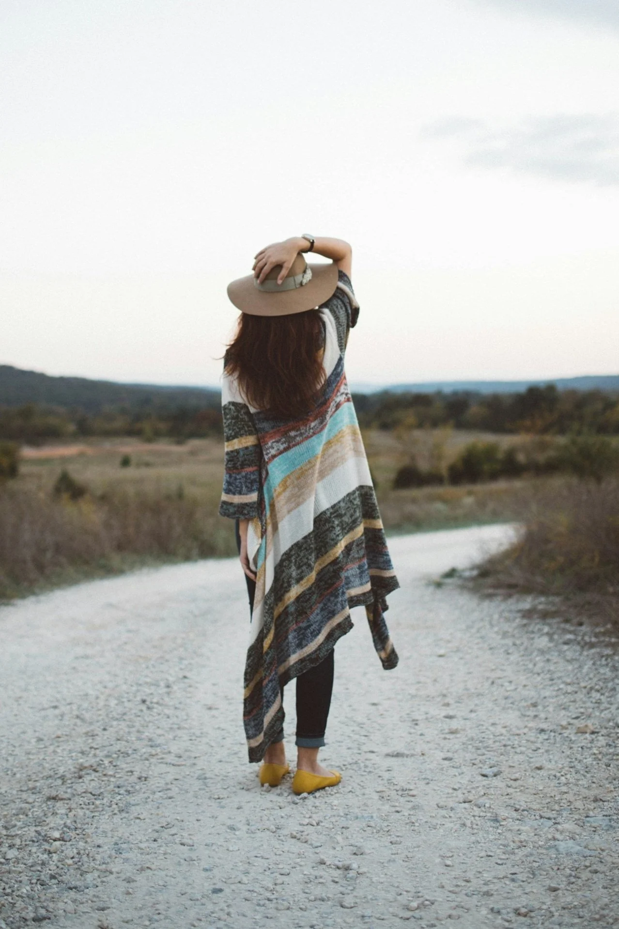 A woman standing on a gravel path in a rural landscape, wearing a wide-brimmed hat, a colorful striped poncho, black pants, and yellow shoes, with her arm raised to hold her hat while looking into the distance.