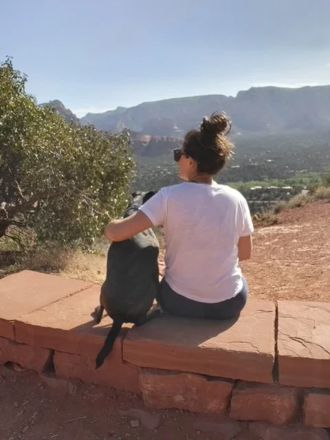 A woman and a dog sitting on a stone ledge looking at a mountainous landscape under a clear sky.