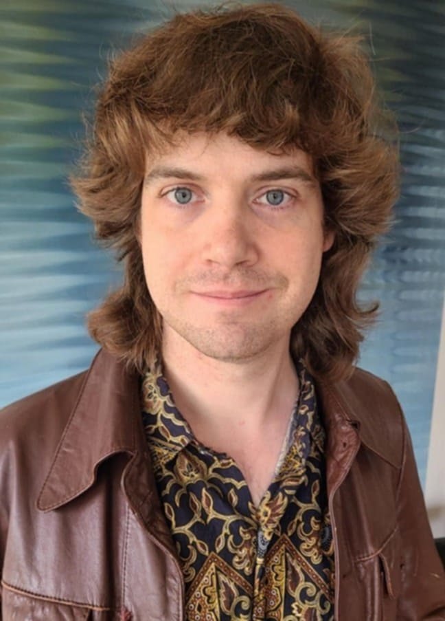 Portrait of Leonard Far with shoulder-length wavy brown hair, blue eyes, wearing a patterned shirt and brown leather jacket, standing in front of a blue abstract background.