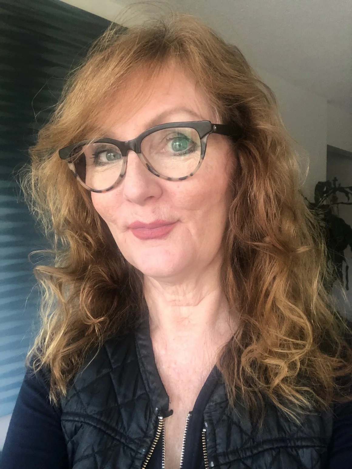 Victoria Clements with red wavy hair wearing large glasses, a black quilted jacket, and a black top, standing indoors.