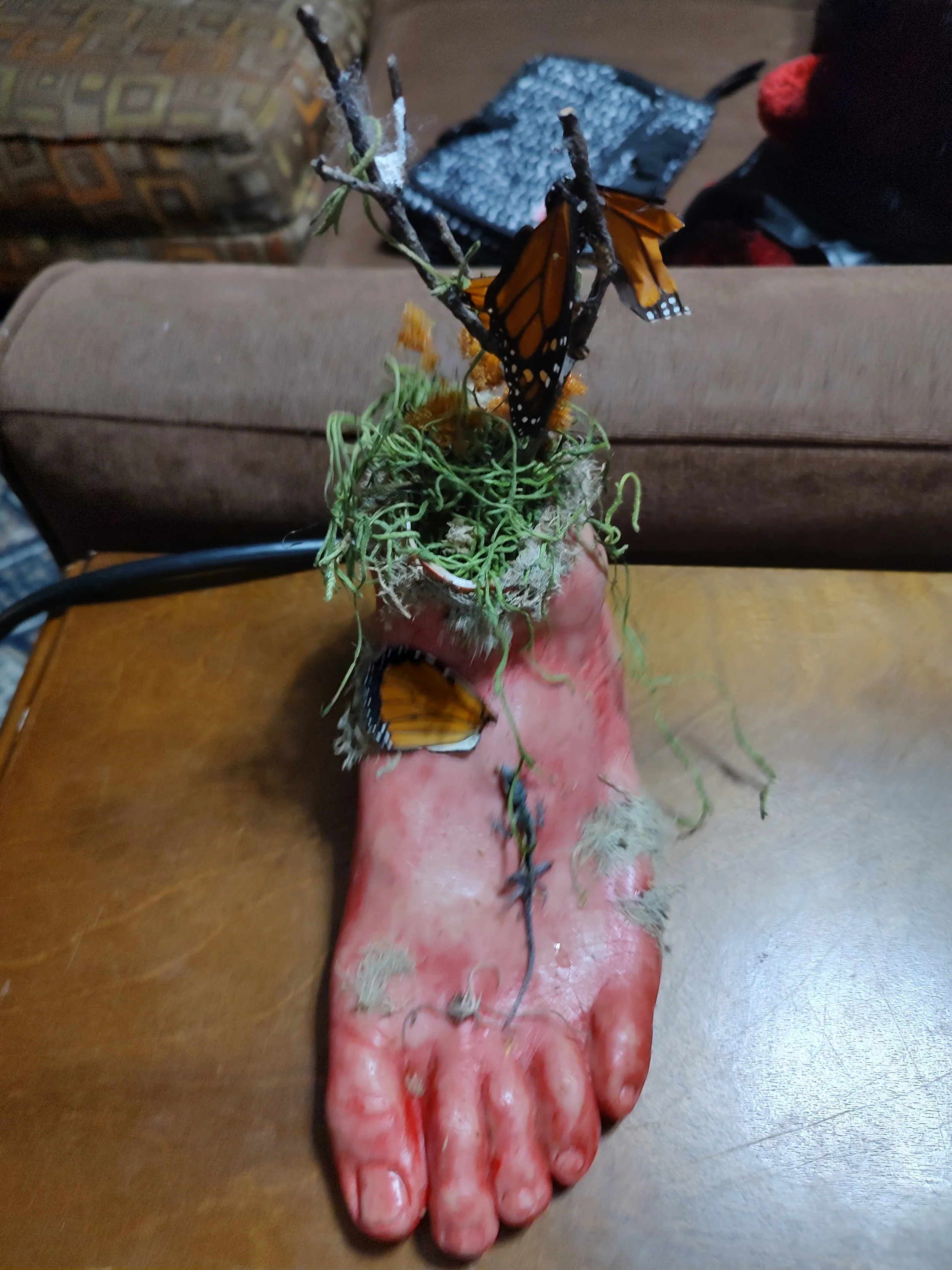 A realistic sculpture of a human foot made of a pinkish material with detailed toes, topped with a miniature garden featuring a branch with Monarch butterflies, moss, and small plants.