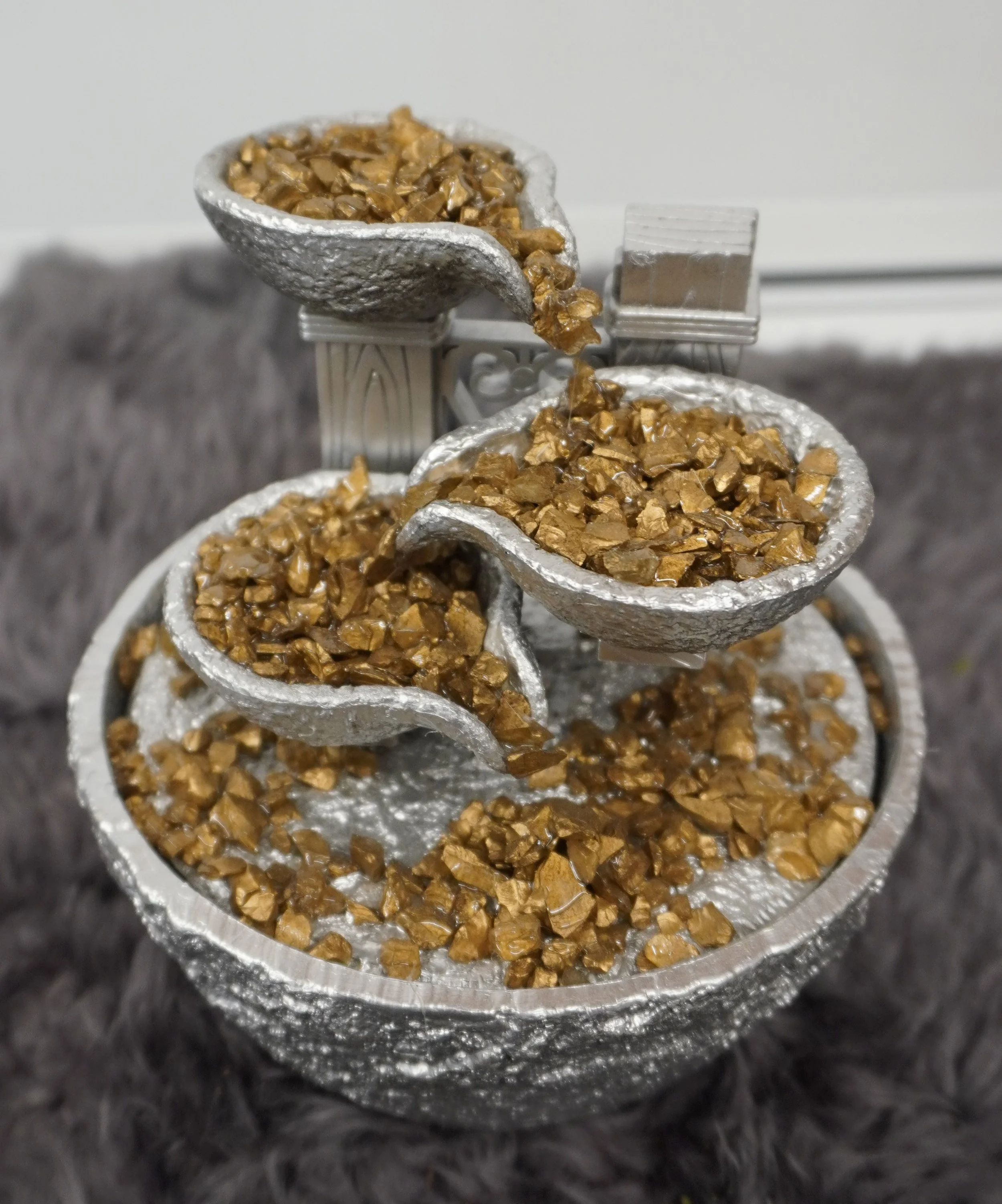 Small three-tiered decorative fountain with gold-colored rocks on a textured gray surface.