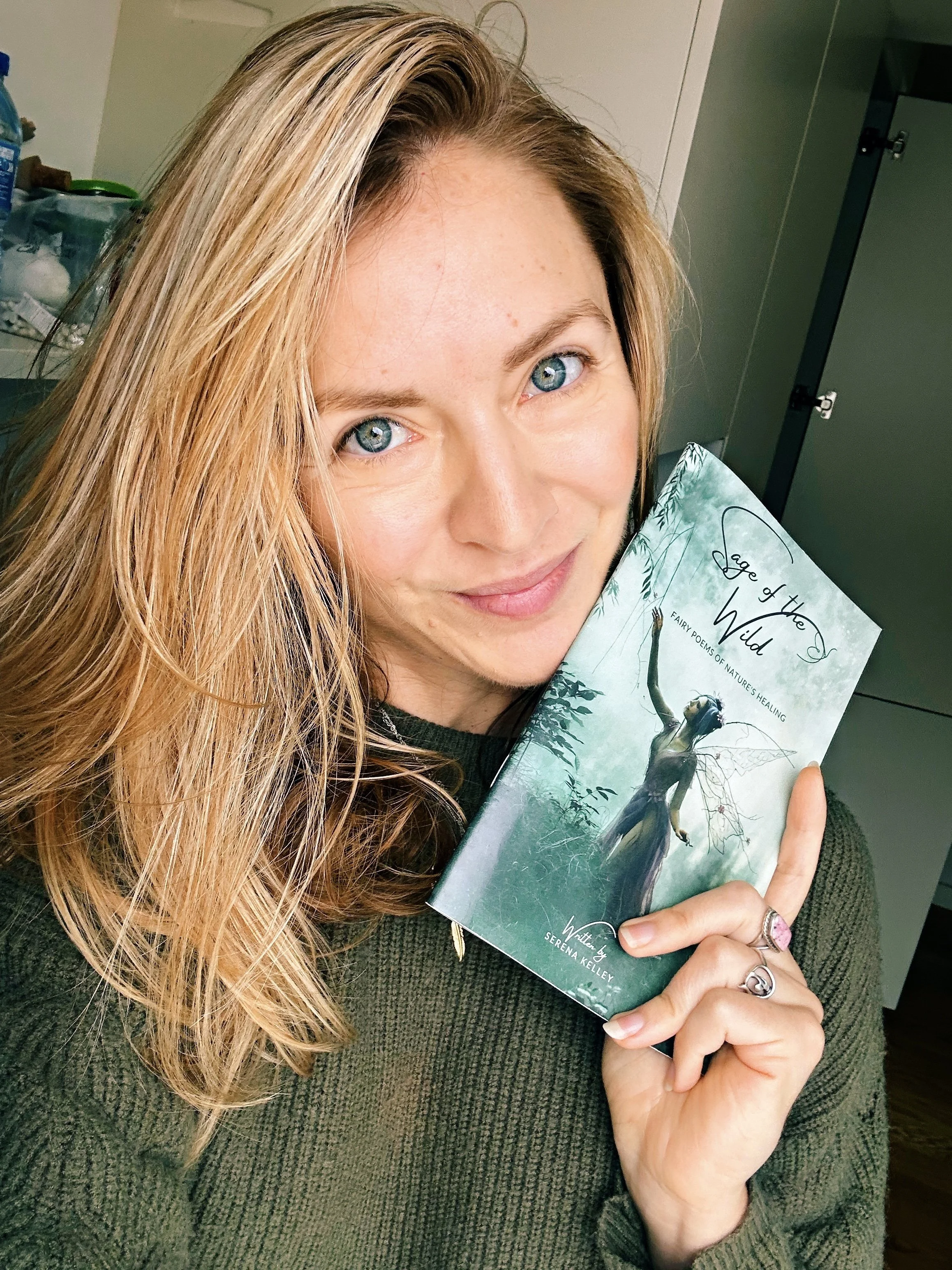 Woman with blonde hair holding a book titled 'Edge of the Wild' in a room with cabinets, smiling at the camera.