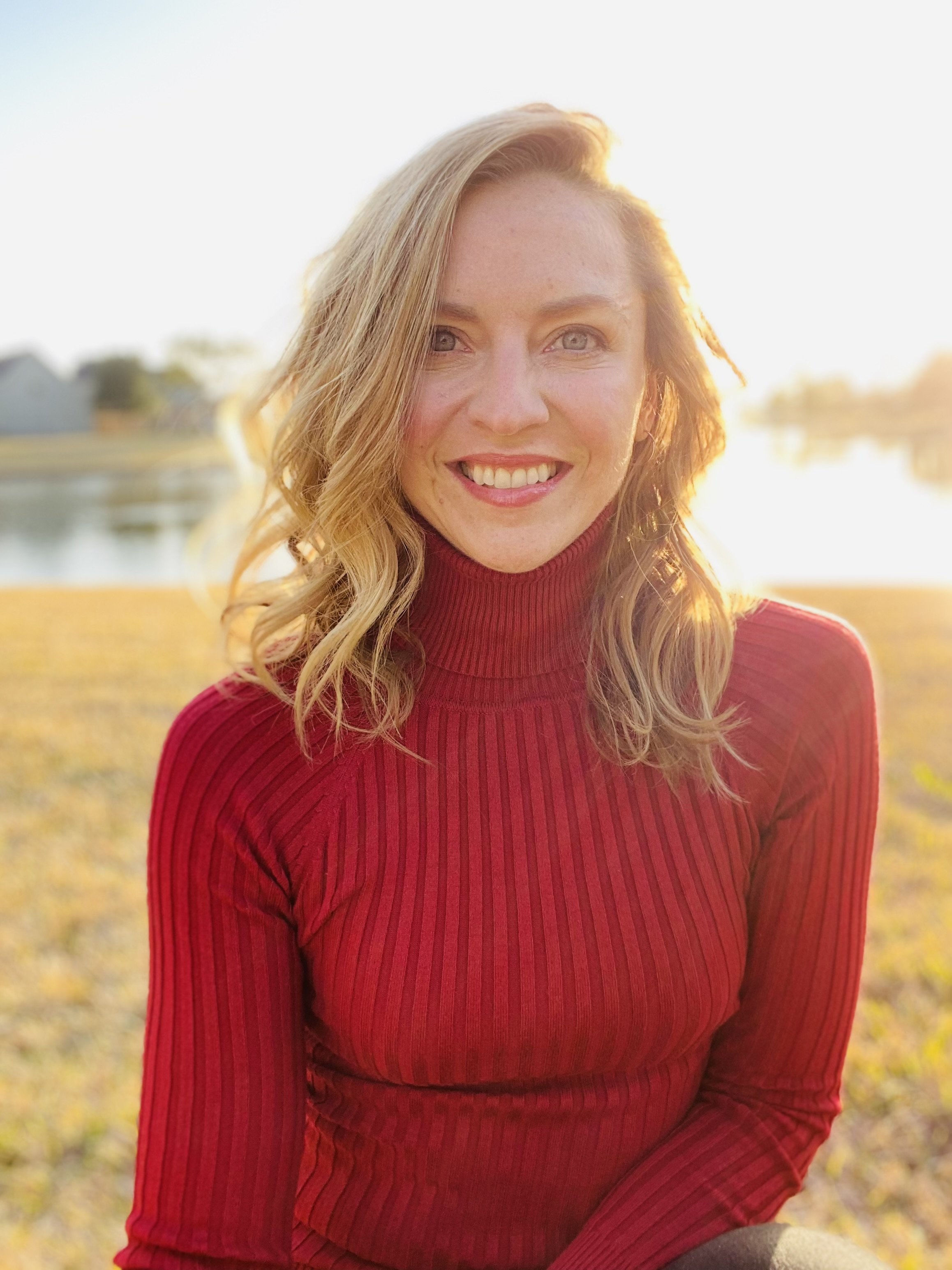 A woman with wavy blonde hair wearing a red turtleneck sweater, smiling outdoors during golden hour with a lake and trees in the background.