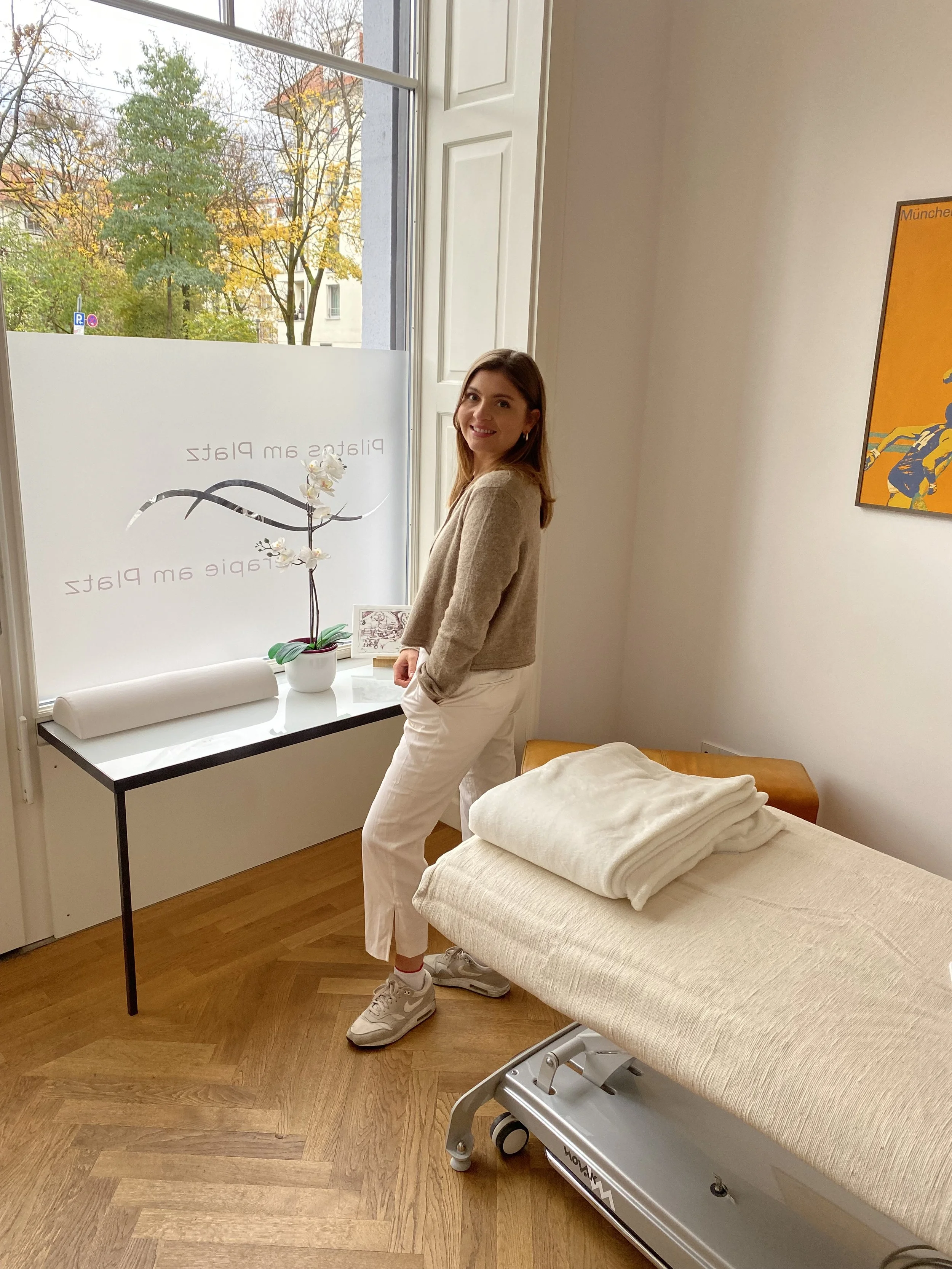 Eine junge Frau steht in einem Raum neben einem Behandlungsbett, trägt beige Kleidung und lächelt. Es gibt eine große Fensterfläche mit Blick nach draußen, auf der herbstliche Bäume zu sehen sind.
