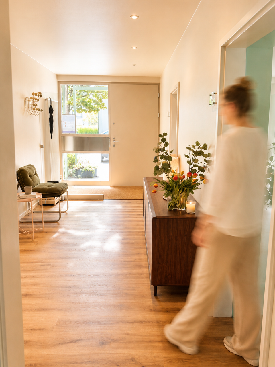 Ein Flur mit Holzboden, einer grünen Sitzgelegenheit, einer Vase mit bunten Tulpen auf einem dunklen Holzschrank und einer jungen Frau, die in einem hellen Outfit vorbeigeht.