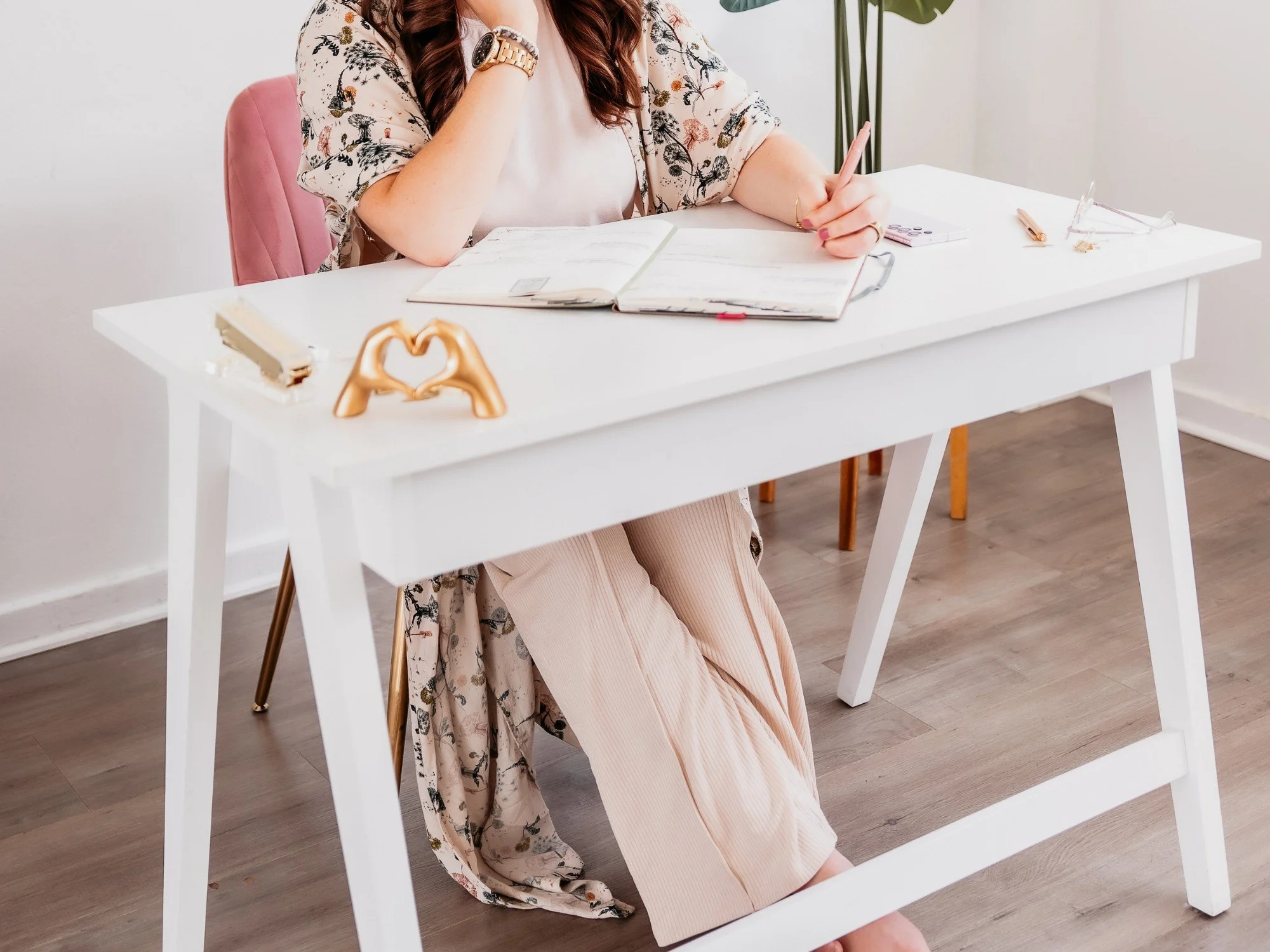 A woman sitting at a white desk, writing in a planner, with a pink chair, a gold heart-shaped sculpture, and a stack of books on the desk. She is dressed in a floral cardigan and beige pants.