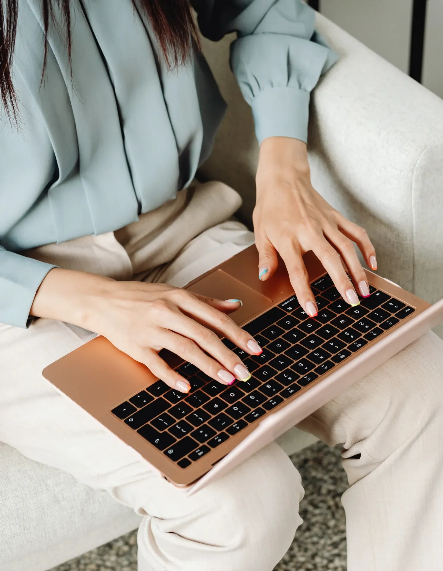 A woman with long hair using a rose gold laptop with a black keyboard, sitting on a light-colored sofa.