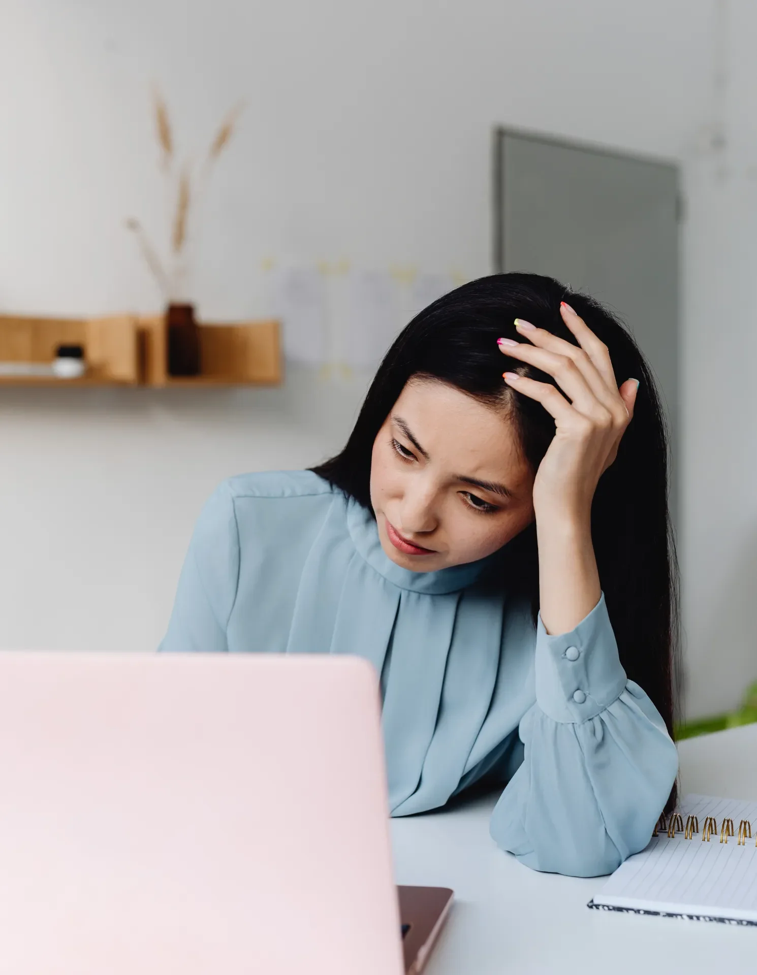 A woman with black hair in a blue shirt sitting at a desk, holding her head in her hand and looking down at her laptop with a worried expression.