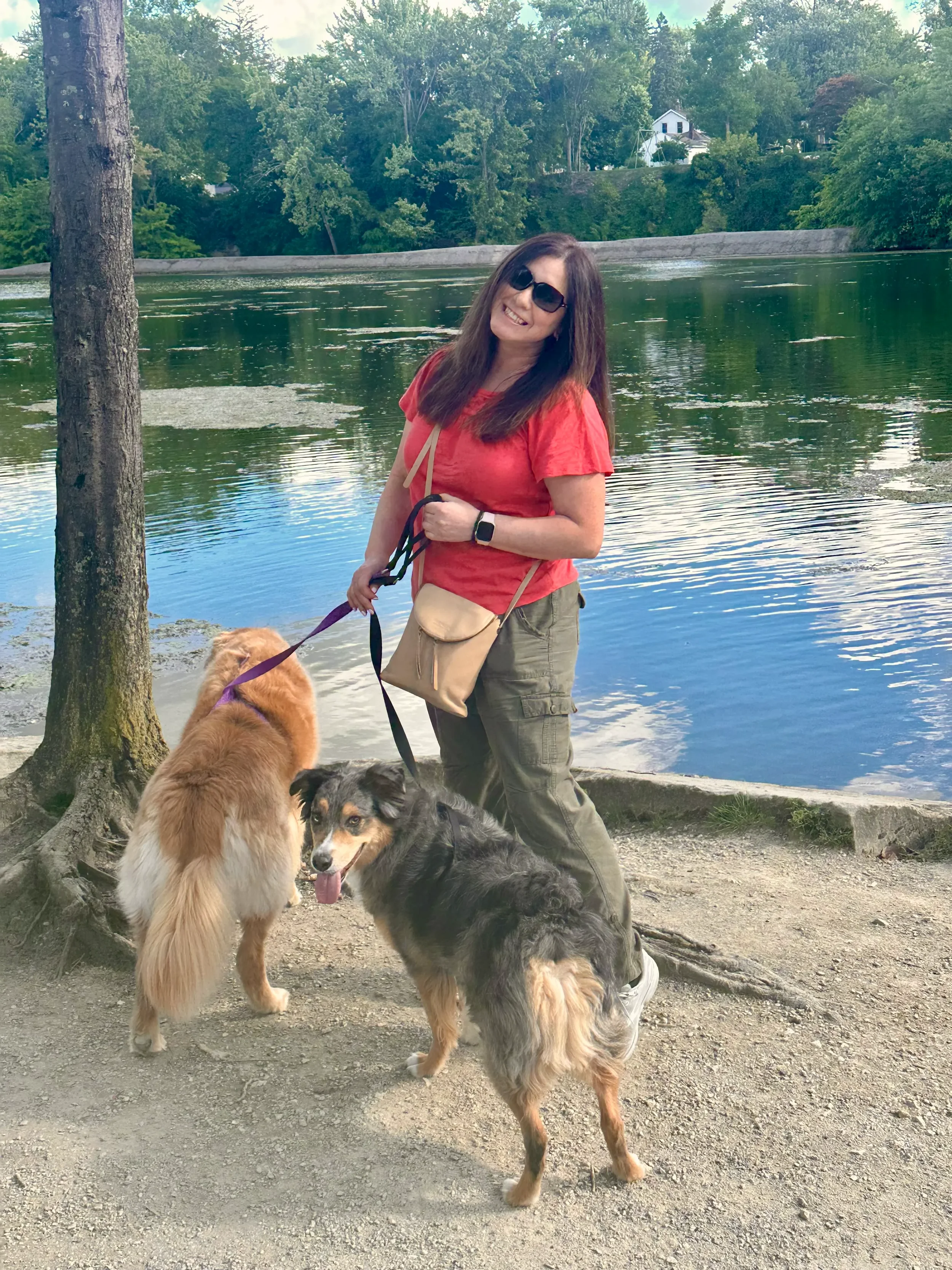 A woman with sunglasses, wearing a red shirt, green cargo pants, and holding two dogs on a leash by a lake with trees and a house in the background.