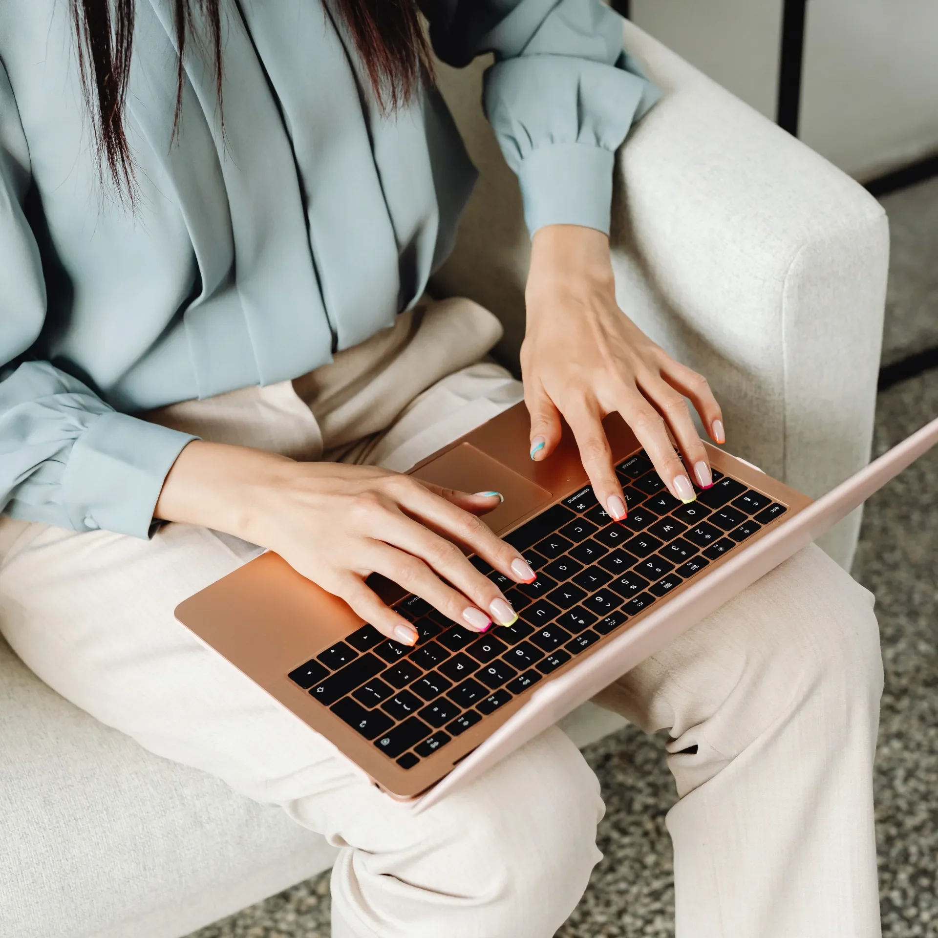 A woman with long hair using a rose gold laptop with a black keyboard, sitting on a light-colored sofa.