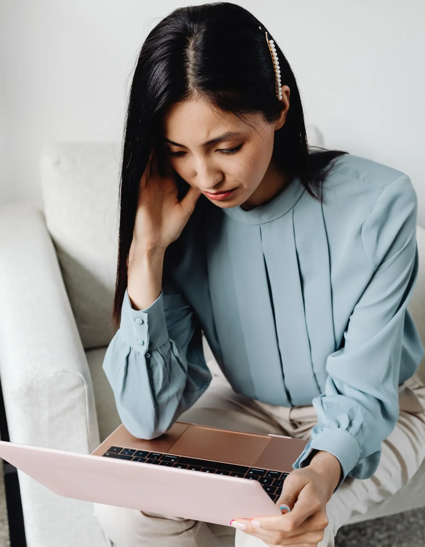 A woman sitting on a light-colored couch using a pink laptop, with her left hand touching her head and wearing a light blue blouse.