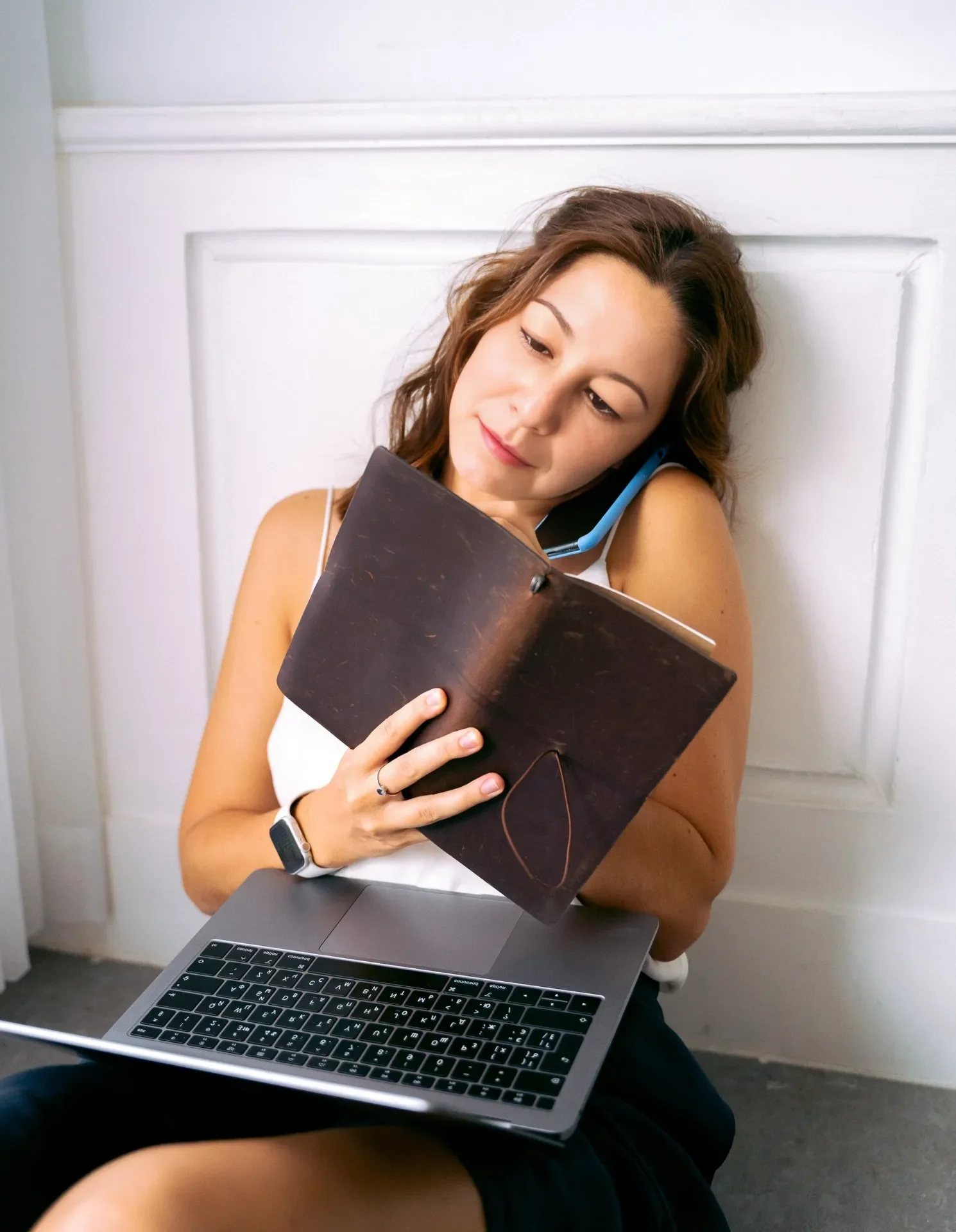Brown haired woman sitting on the phone, while writing in a notebook, with a laptop on her lap.