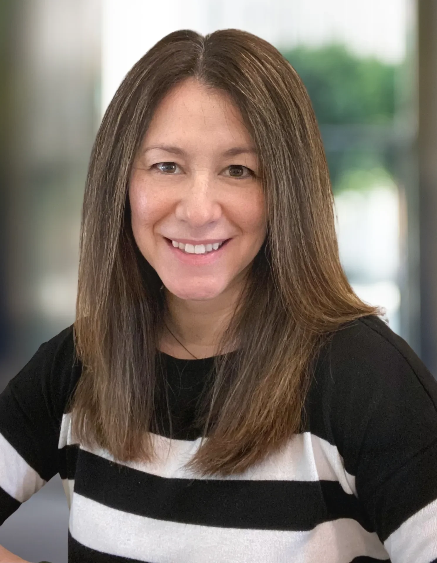 Tomi Lucien, a woman with long, straight brown hair and light skin smiling at the camera. She is wearing a black and white striped top with a black sleeve. The background is blurred with natural light.