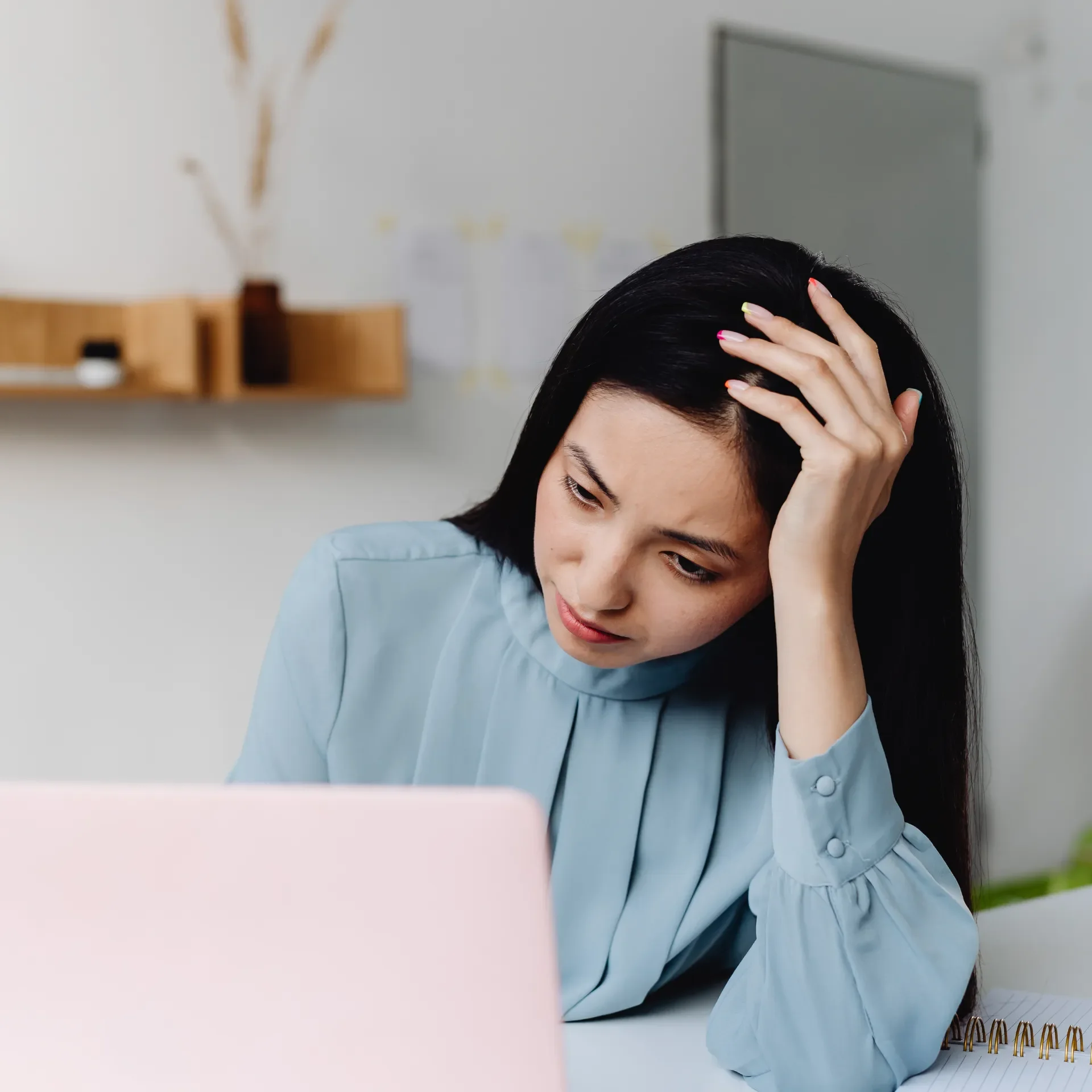 A woman with black hair in a blue shirt sitting at a desk, holding her head in her hand and looking down at her laptop with a worried expression.