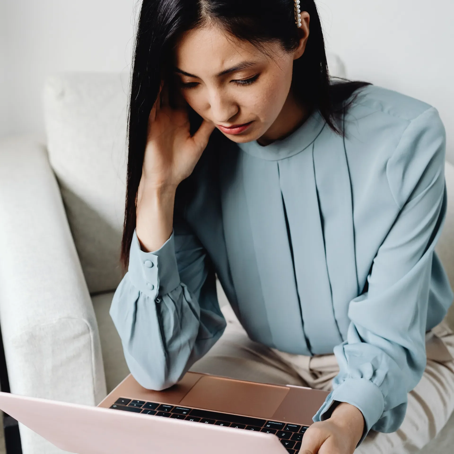 A woman sitting on a light-colored couch using a pink laptop, with her left hand touching her head and wearing a light blue blouse.