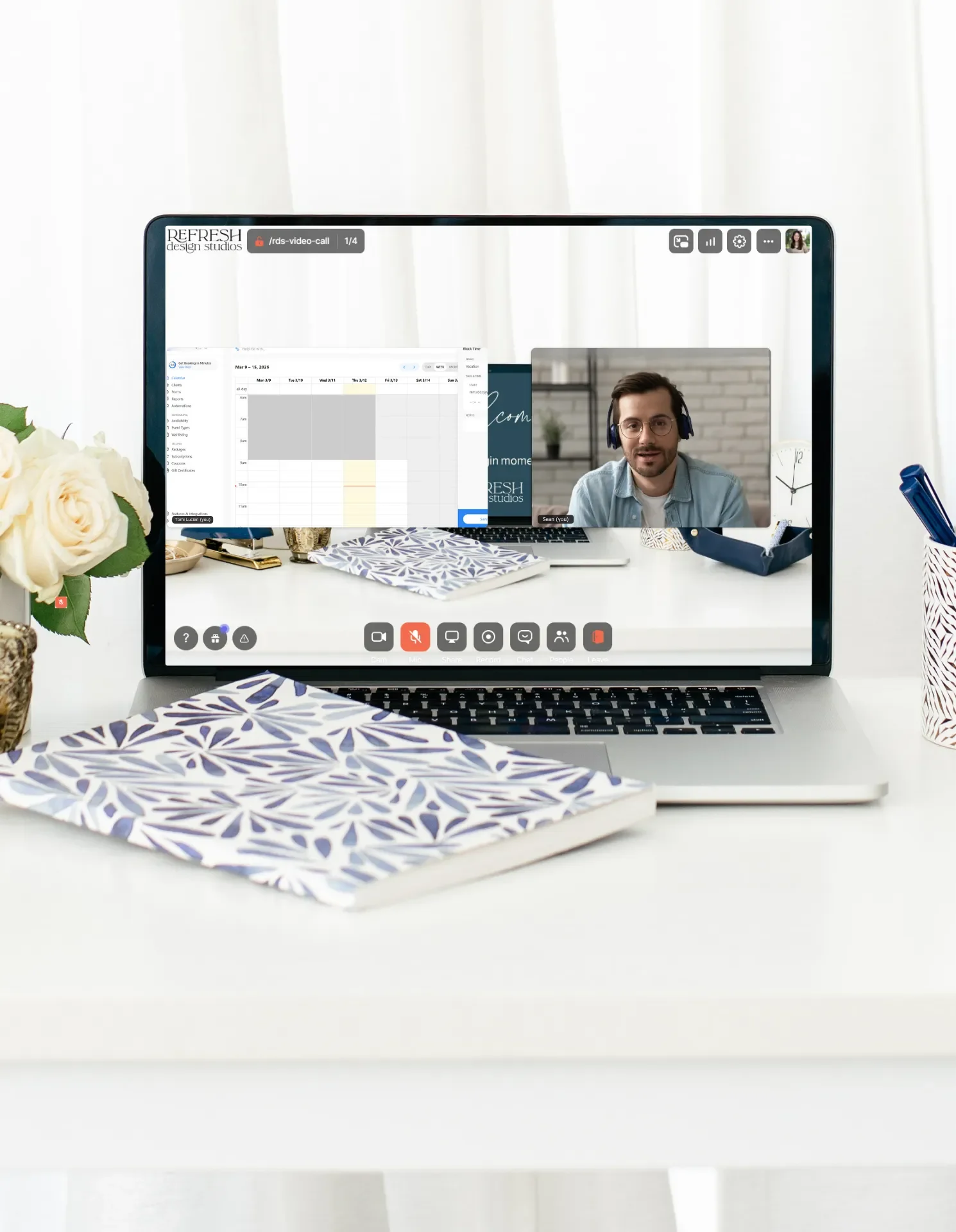 A laptop on a desk with a Zoom-type meeting on the screen and a blue palm leaf notebook closed across the keyboard
