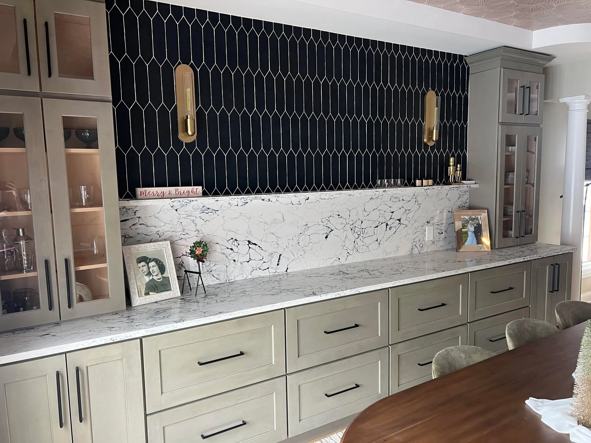 A kitchen or dining area with light gray cabinetry, a marble countertop with white and black veining, and a matching marble backsplash. Decorative items include framed photographs, a small plant, and a sign reading 'Merry & Bright.' The wall above features black textured tiles with gold wall sconces.