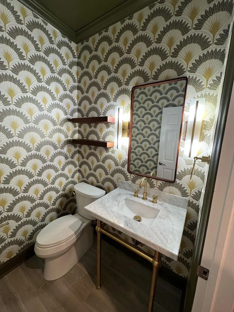 A small bathroom with patterned wallpaper featuring fan-shaped designs in gray, black, and gold. It has a white toilet and a marble vanity with a gold faucet and fixtures. A rectangular mirror with a thin gold frame hangs above the vanity. Two vertical wall sconces with modern white shades are mounted on either side of the mirror. There are two dark wood open shelves mounted on the wallpapered wall to the left of the mirror.