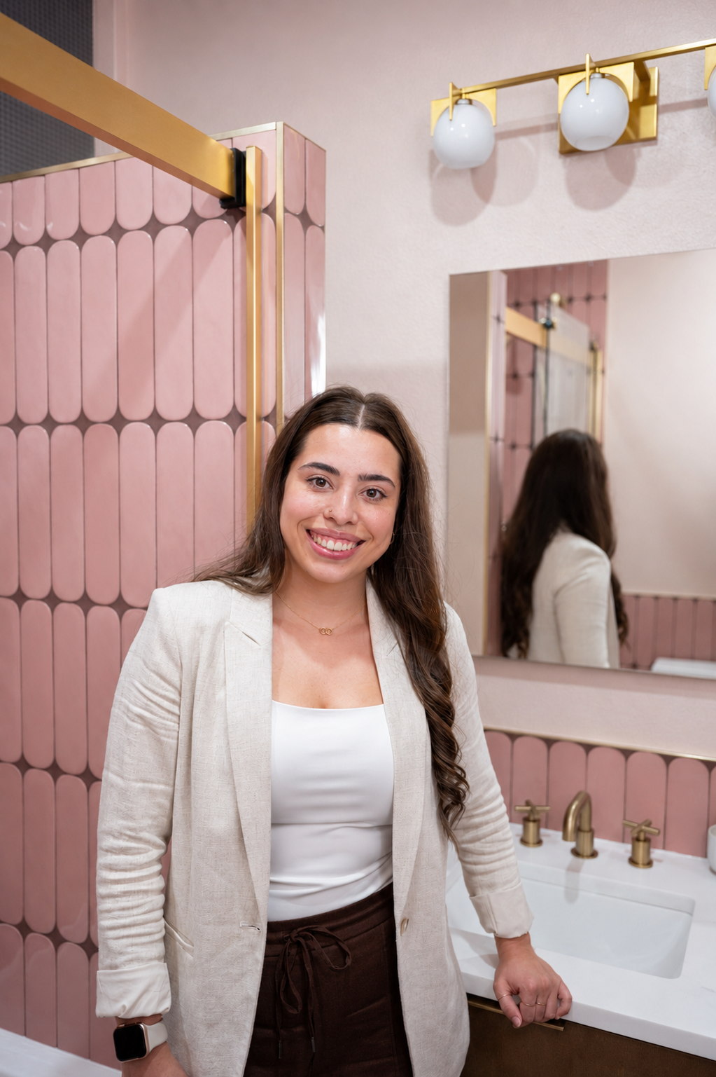 A woman with long brown hair smiling in a modern pink and gold bathroom, standing next to a sink, with a mirror reflecting her back.