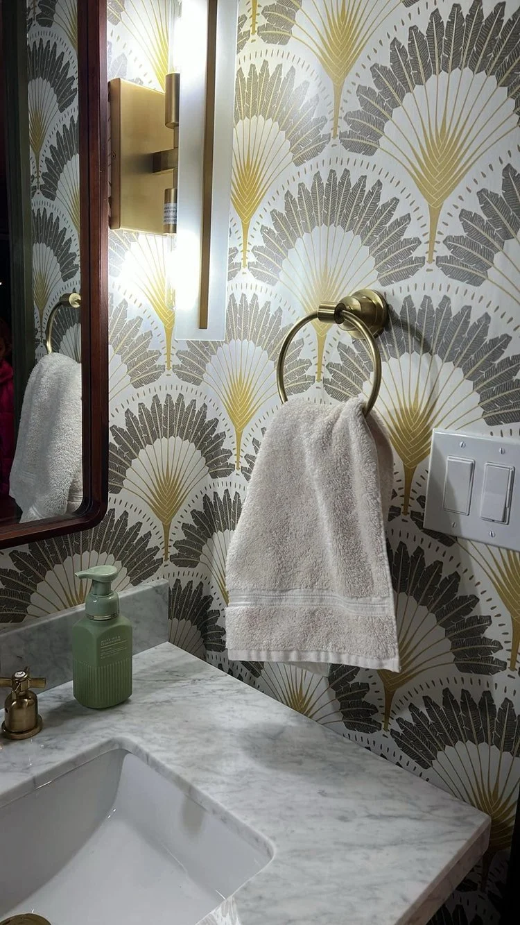 A bathroom sink with a marble countertop, a green liquid soap dispenser, and a gold faucet. A towel hangs on a gold ring next to a distinctive gold wall sconce and a mirror with a dark wood frame. The wallpaper features a pattern of gold and dark gray palm leaves.