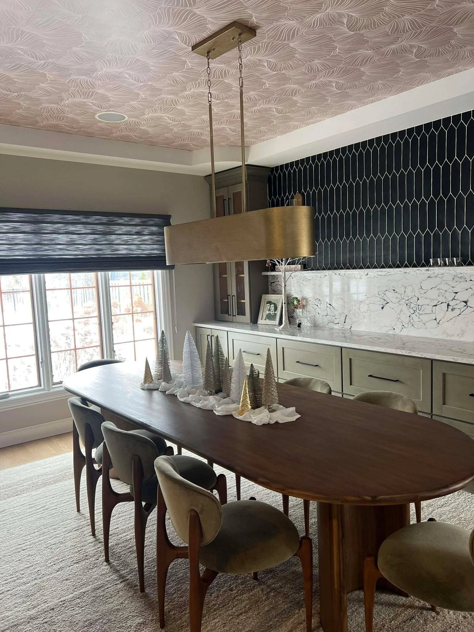 A dining room with a wooden table surrounded by eight chairs, a long white sideboard with black handles, a marble backsplash, a black tile wall, a window with gray curtains, and holiday decorations including a centerpiece of small Christmas trees on the table.