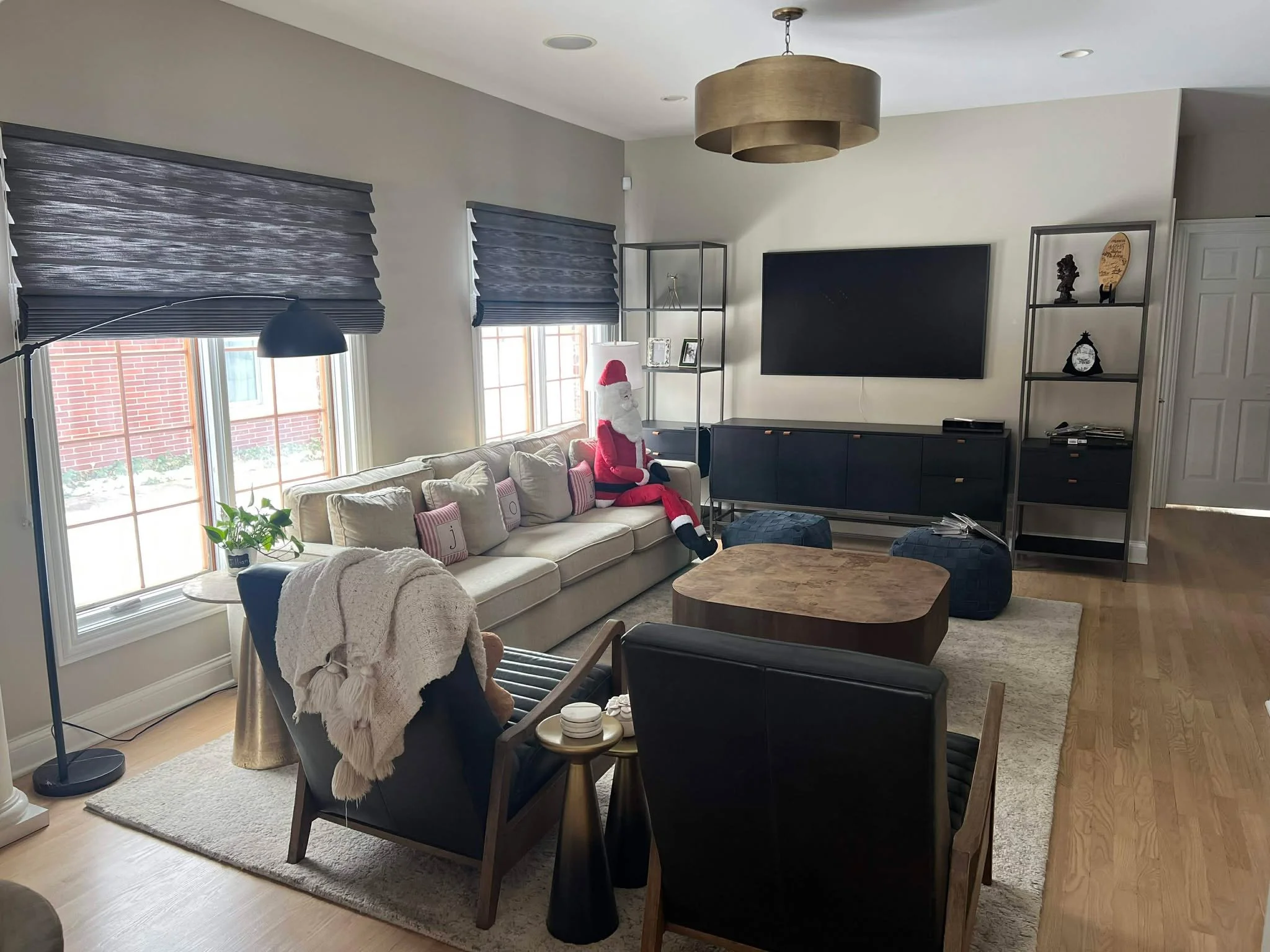 A living room with a beige sofa decorated with holiday pillows, a Santa Claus figure seated on it, and a mix of modern and traditional furniture. Multiple windows with blue shades, a black TV on the wall, open shelving with decorative items, and a large beige drum-shaped ceiling light.