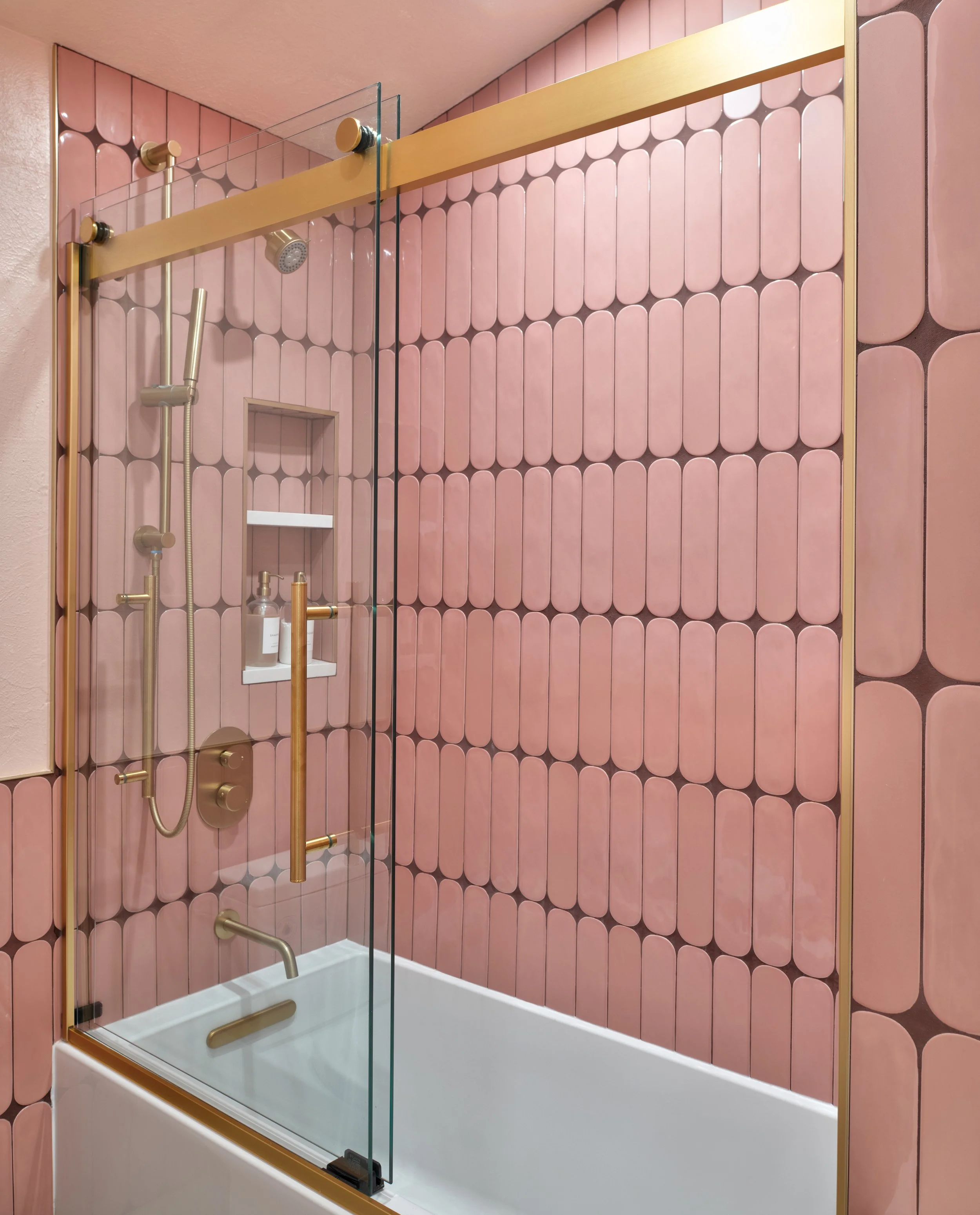 Pink tiled bathroom shower with gold fixtures, a glass shower door and niche for toiletries.
