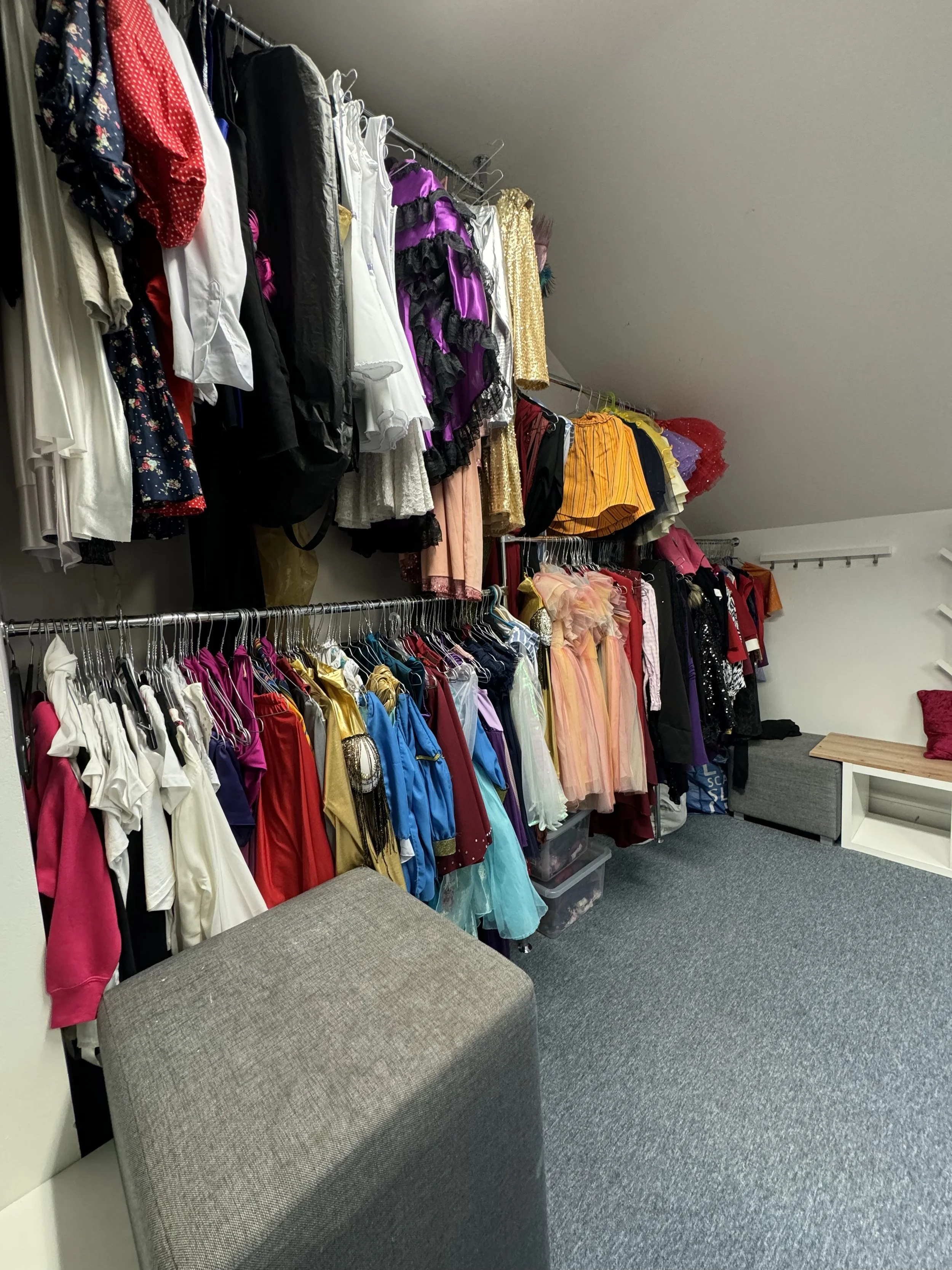 Closet with two rows of hanging dresses and clothing, a small shelf with a pillow, and a gray ottoman.