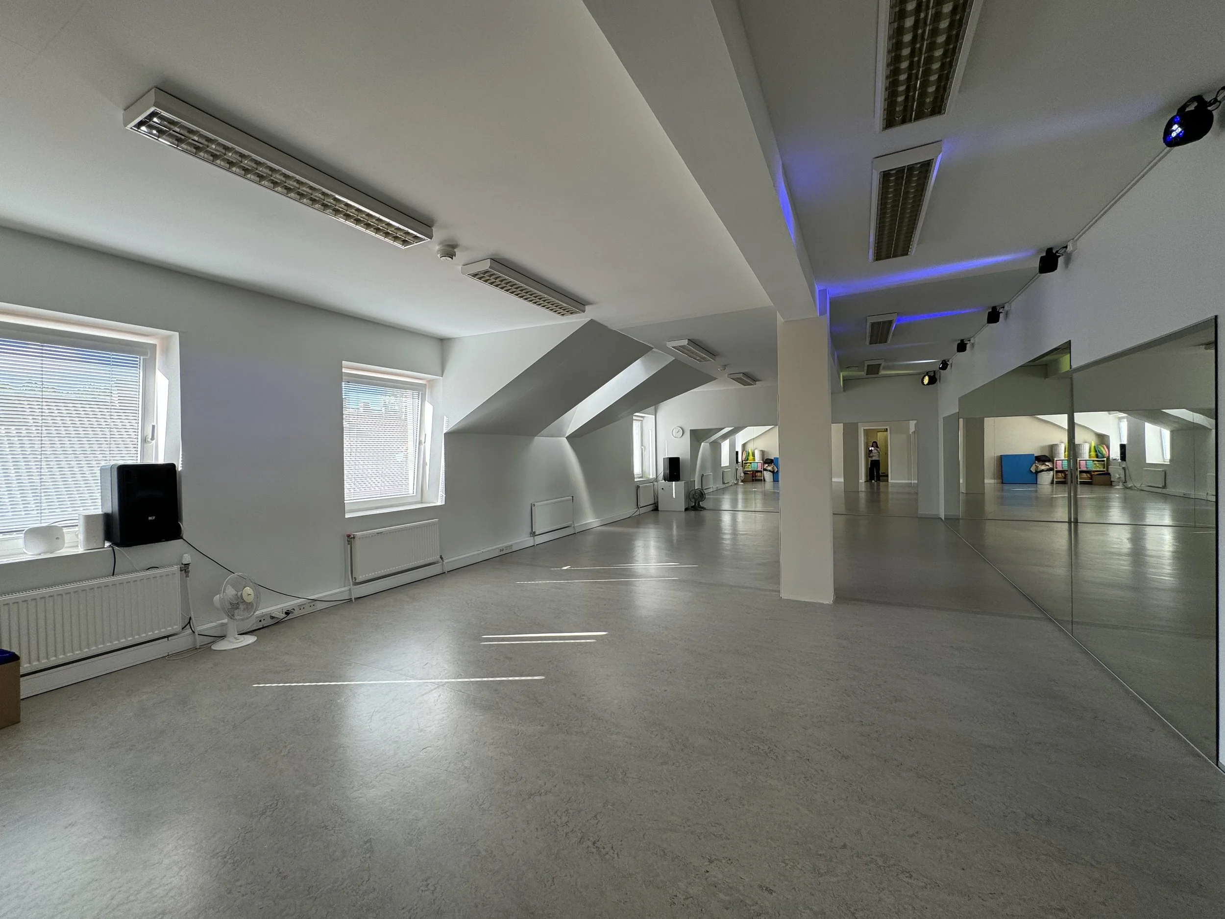 Empty dance studio with light-colored flooring, white walls, large mirrors on one wall, and Windows with blinds, some equipment and a person in the background.