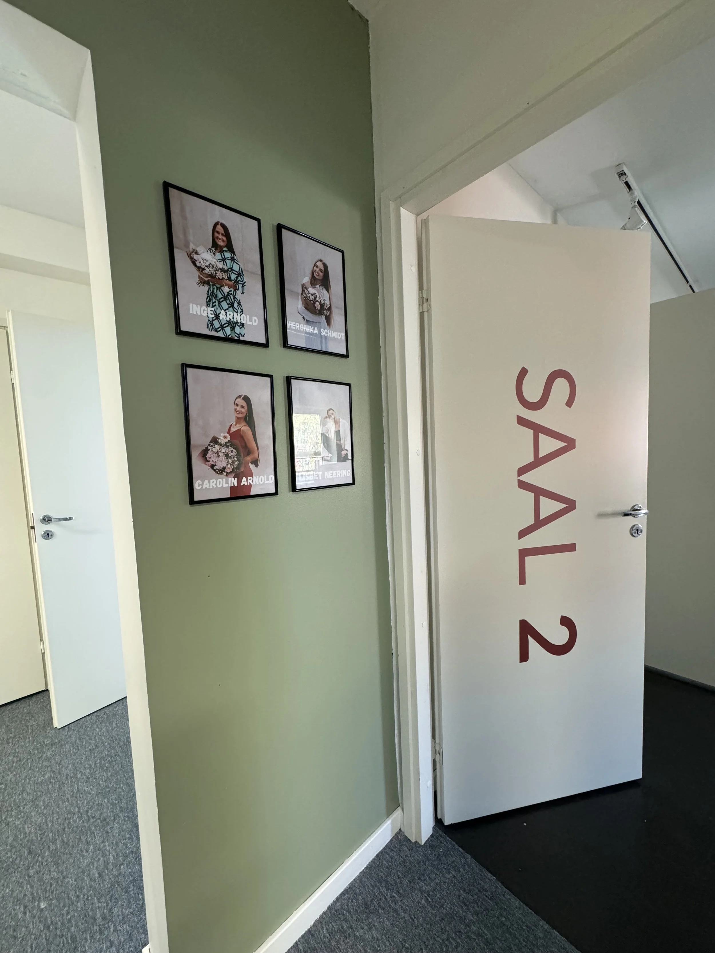 Photo of an office hallway with framed pictures of four women on a green wall. To the right, a door marked 'SAAL 2' is partially open.