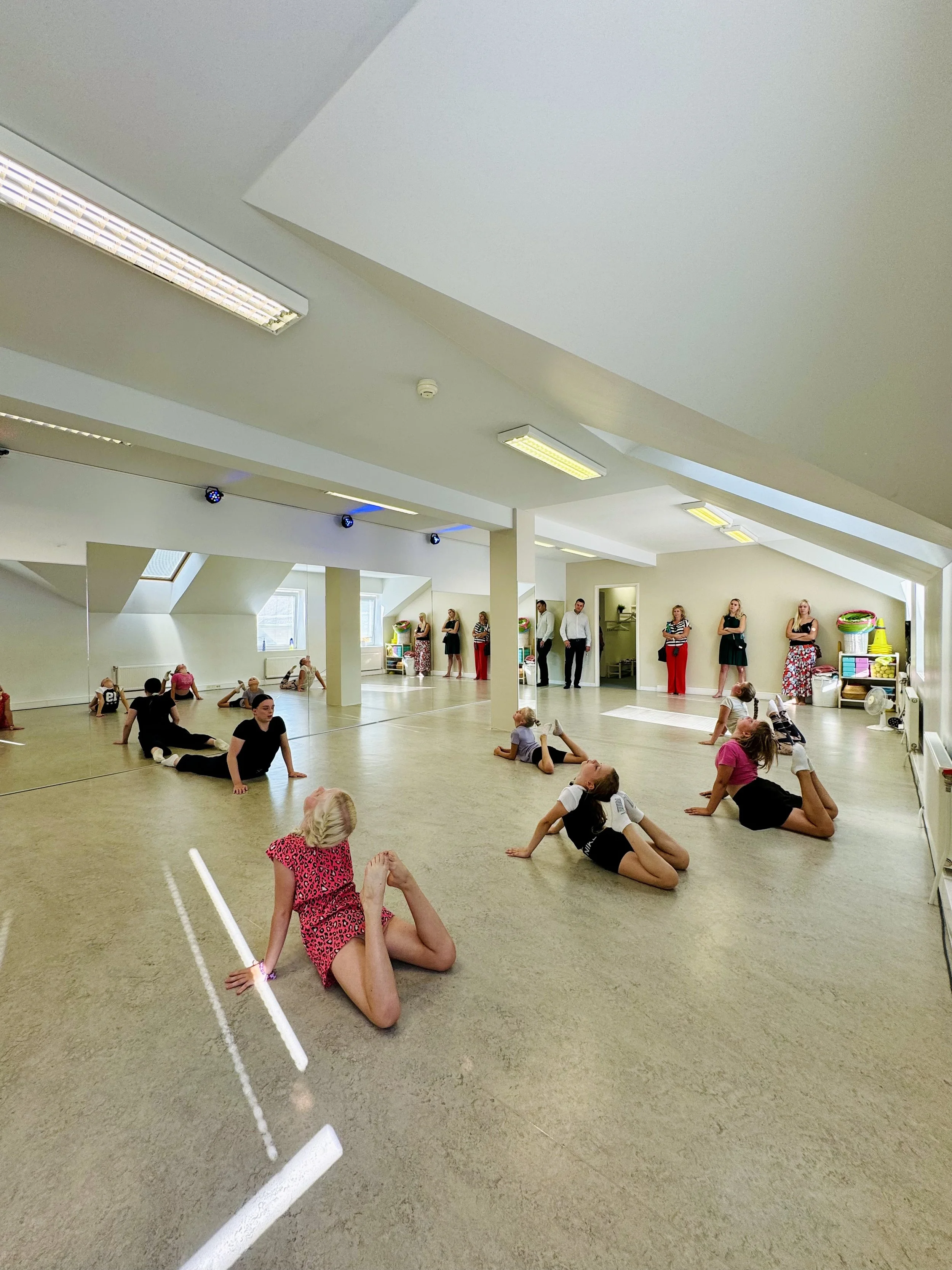 Children participating in a dance or gymnastics class in a spacious, well-lit room with a sloped ceiling, mirrors, and a group of women watching near the back of the room.