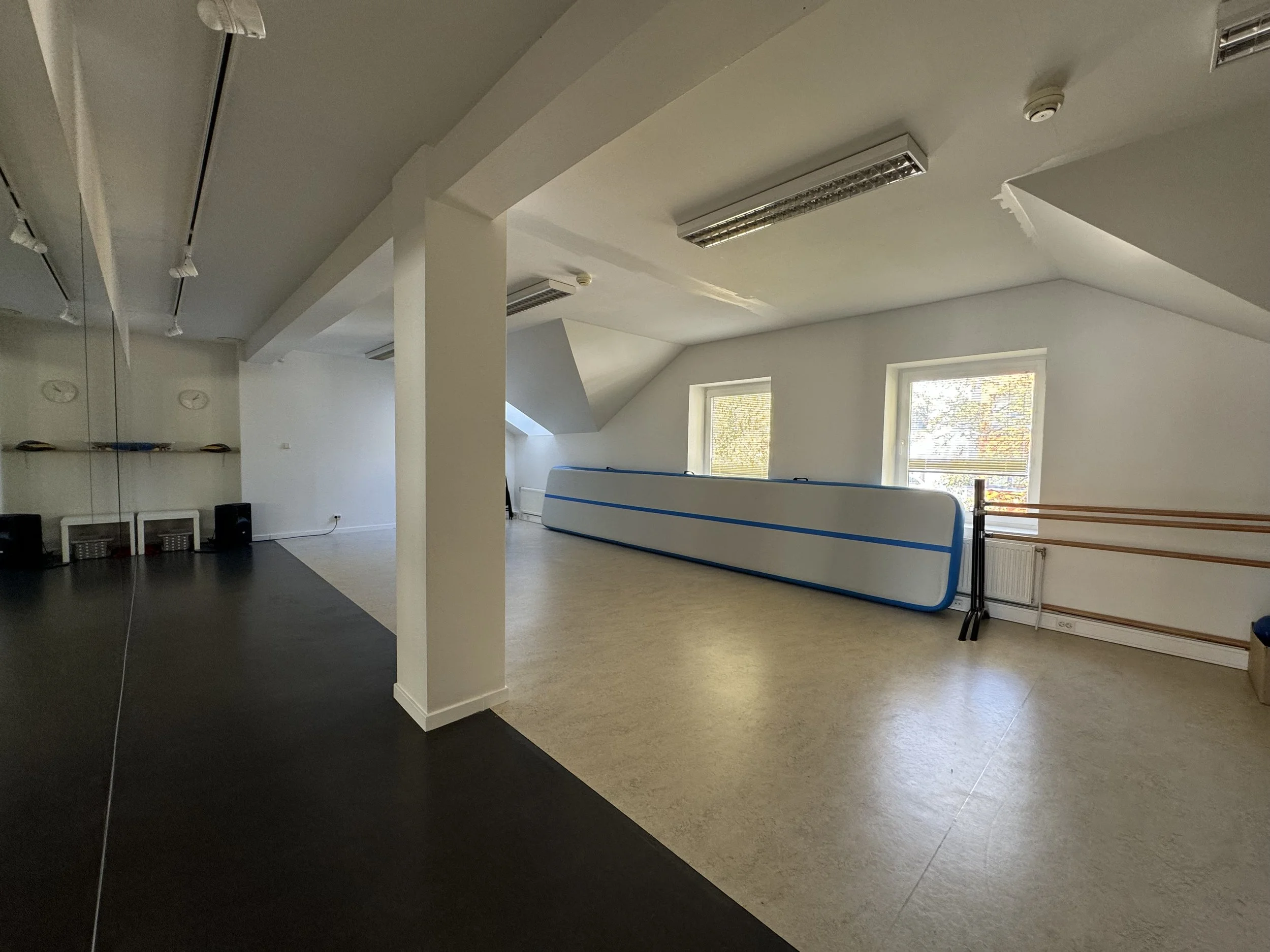 Empty dance studio with large windows, mirrored wall, ballet barres, and a folded gymnastics mat.