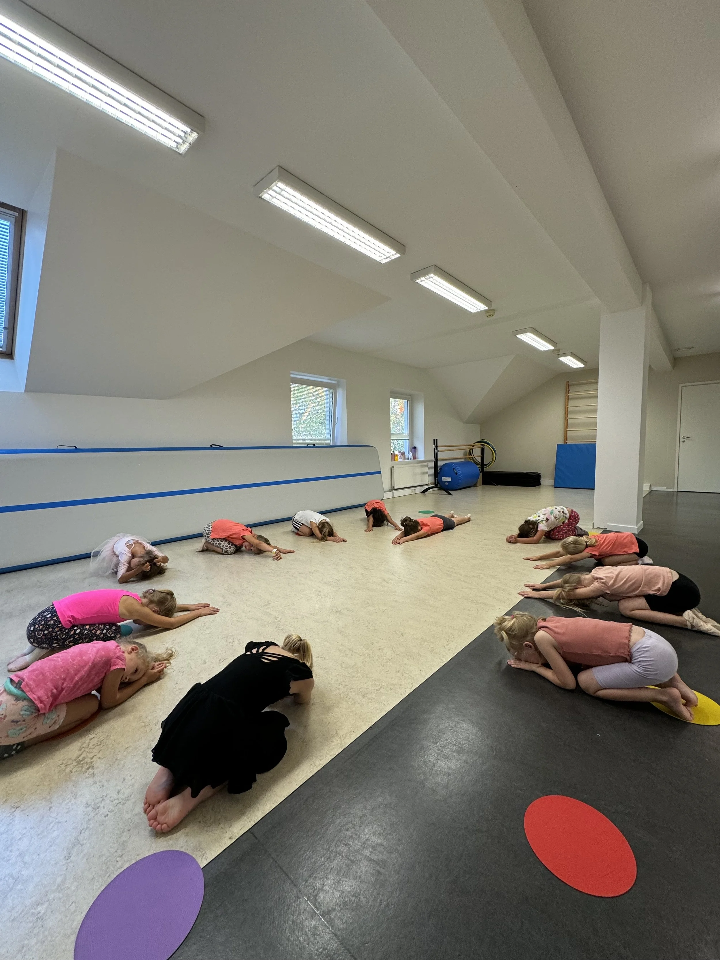 Children participating in a yoga class in a bright, spacious studio, practicing a kneeling forward bend pose.