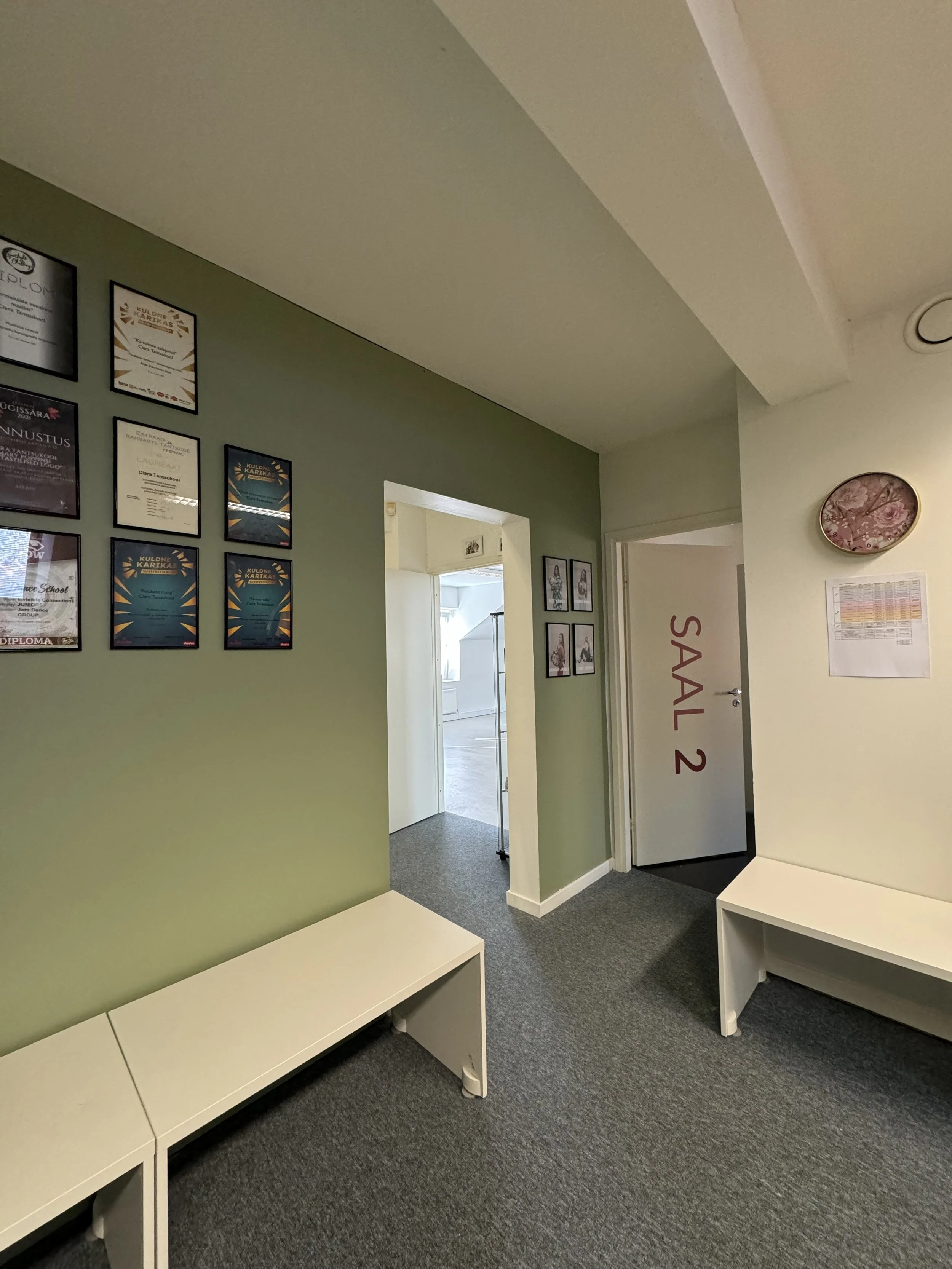 Interior of a waiting area with green and cream walls, framed certificates, pictures, white benches, and a door labeled 'Saal 2'.