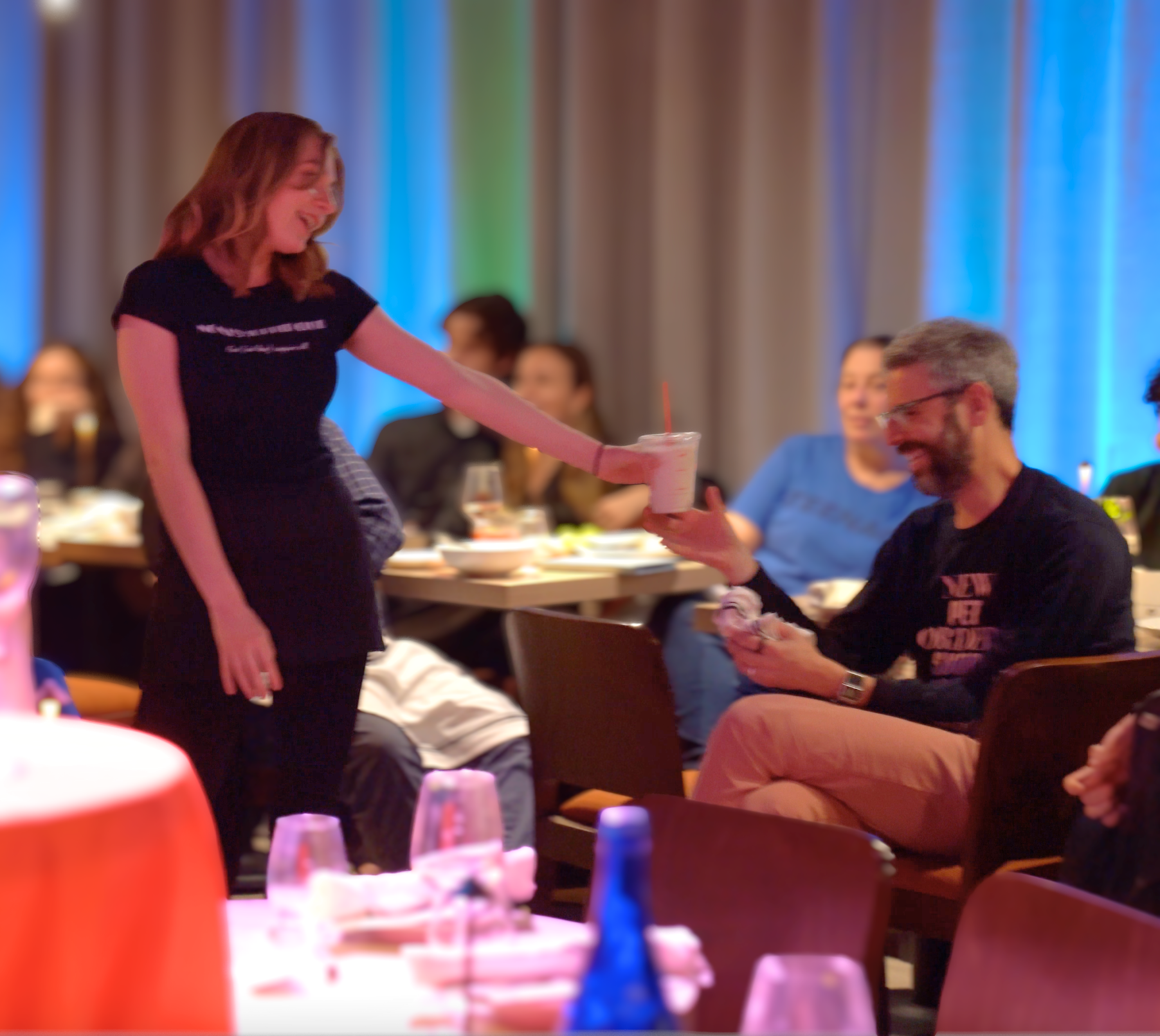 A woman in a black shirt is handing a man a drink at a social gathering. The man is smiling as he receives the drink. Other people are seated at tables in the background, enjoying the event.