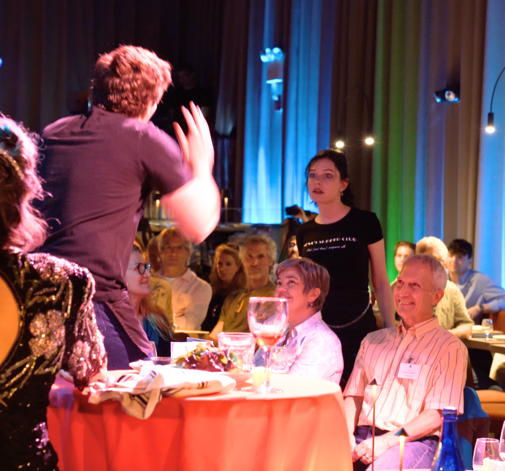 A group of people at a social event, with a person standing and speaking to the seated audience. The audience appears attentive, with some smiling and others looking surprised. The setting looks like a dinner or banquet with tables, drinks, and color