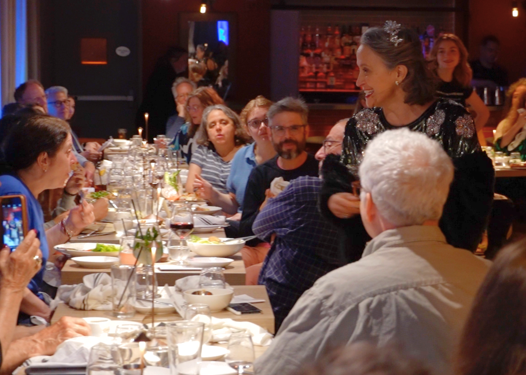 A woman in a black, embellished dress is standing and smiling at a group of people seated at a long dining table in a restaurant. The table is set with plates, glasses, and salads, and most people are looking at her. The setting appears to be a livel