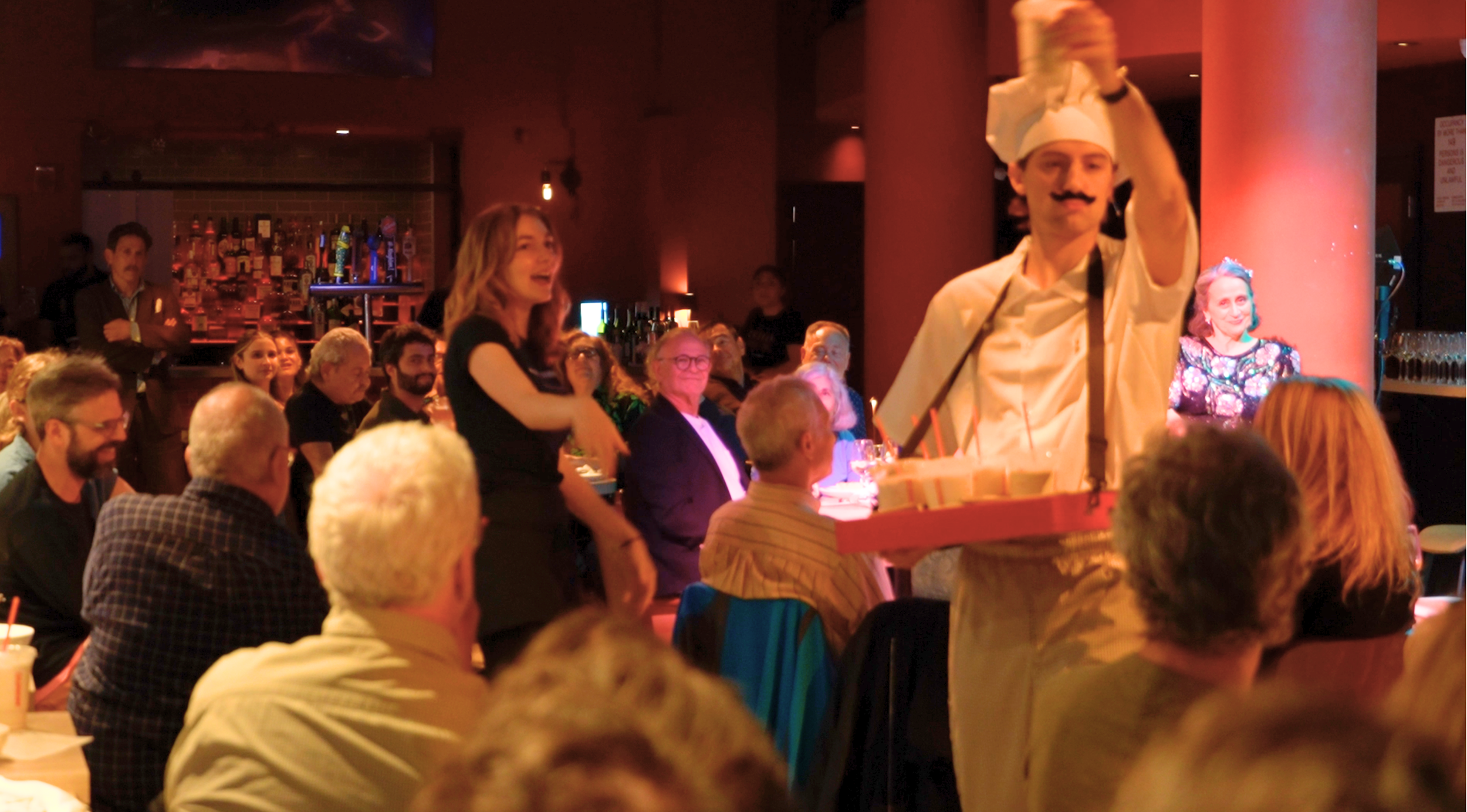Chef with Mustache serving drinks to audience at a lively event in a dimly lit bar or restaurant.