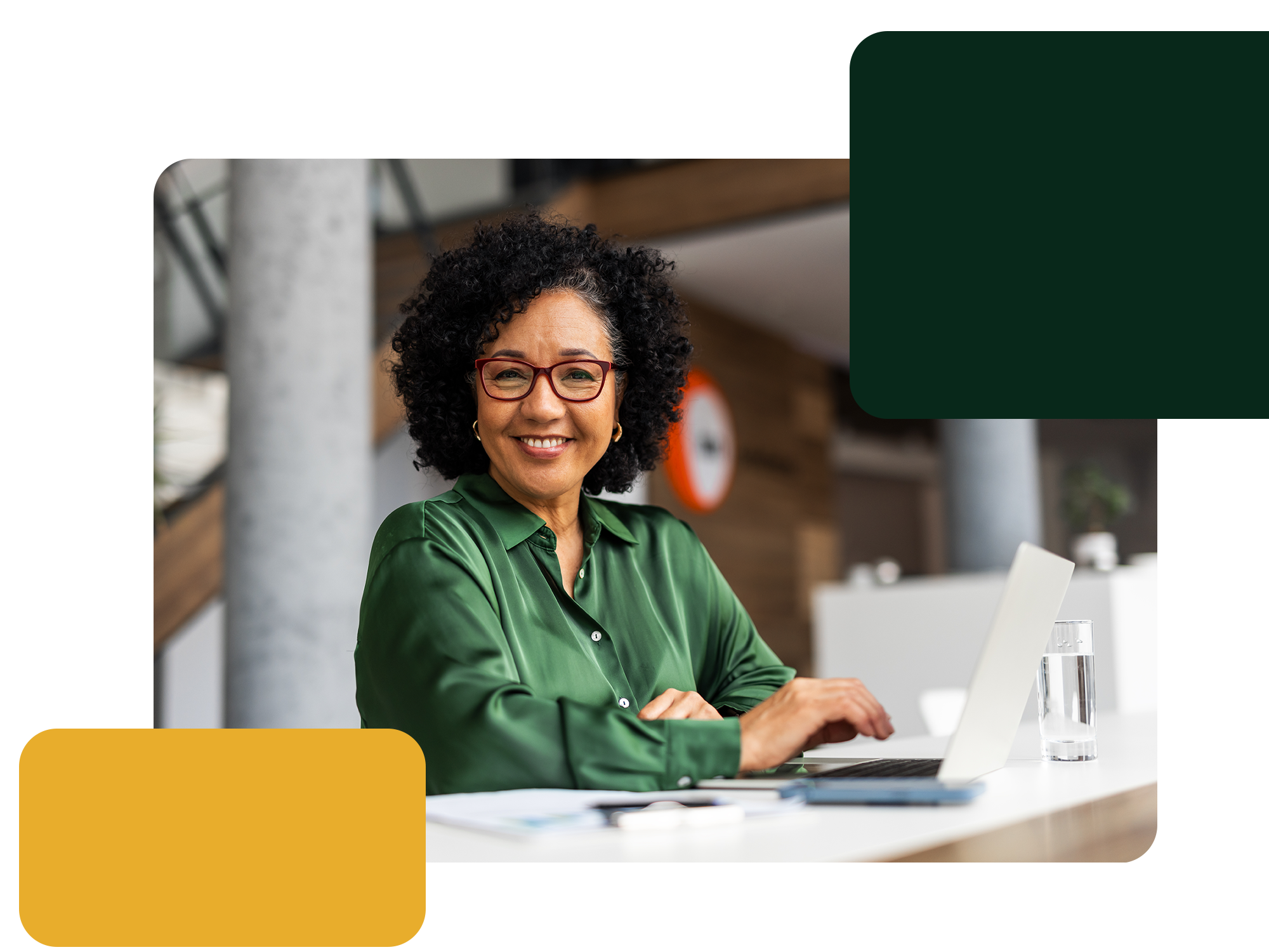 Smiling woman with curly hair and red glasses wearing a green blouse sitting at a desk with a laptop and a glass of water, in a modern office setting.