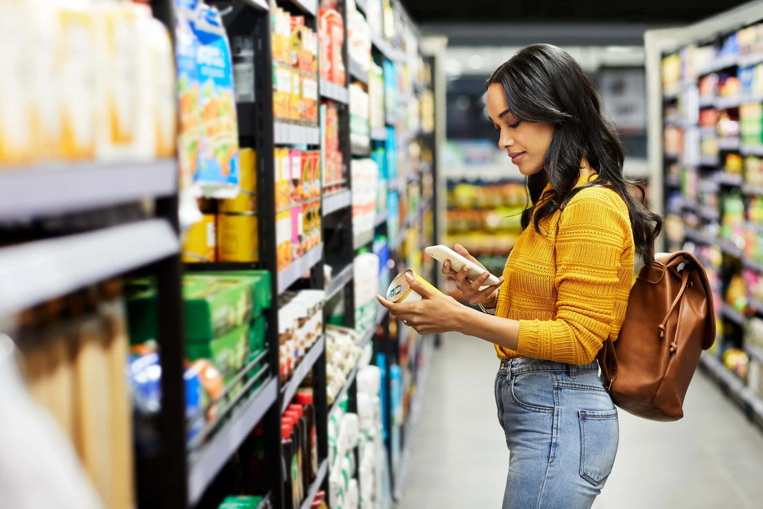 Young woman shopping in a grocery store, holding a can and using her phone.